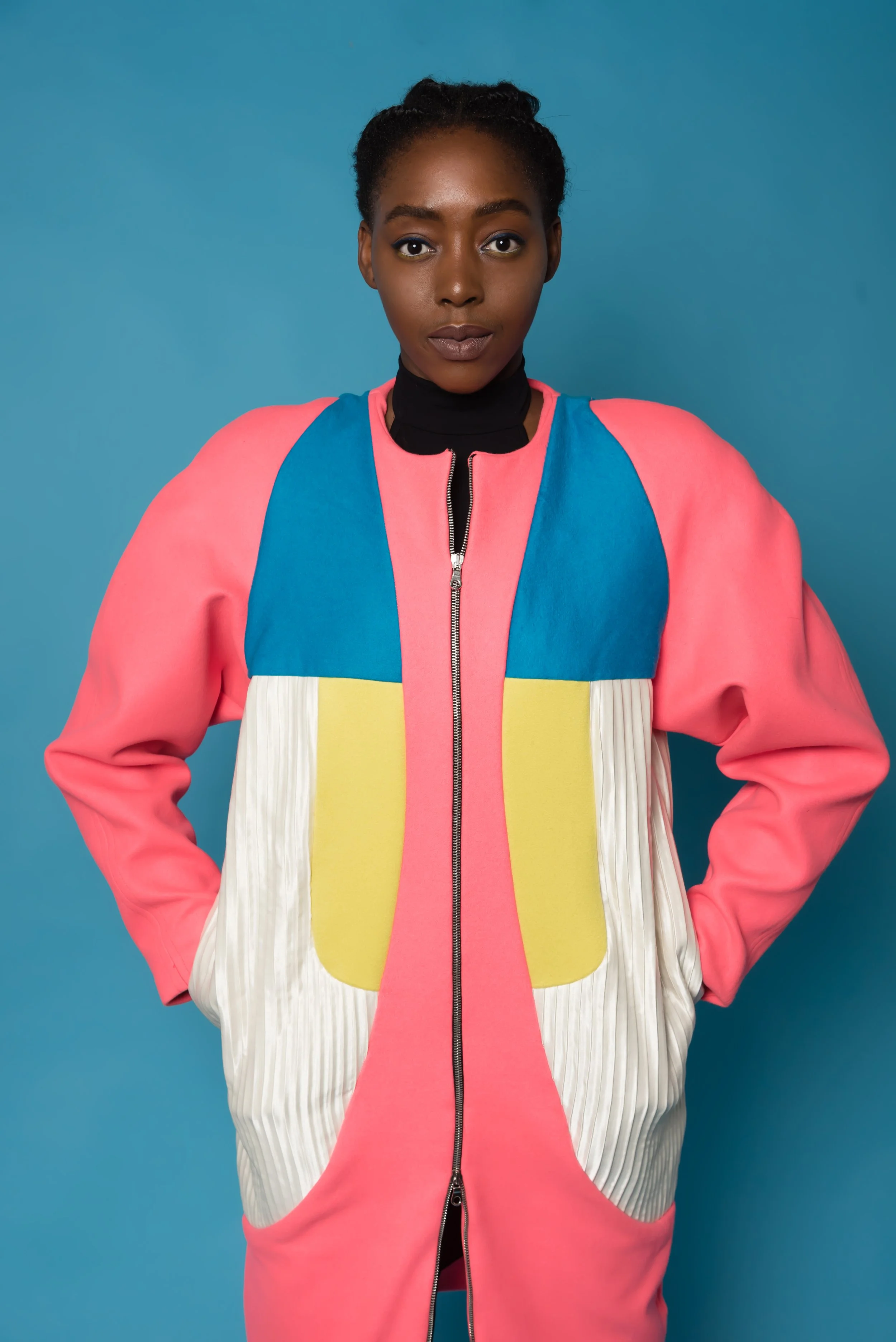 A woman wearing a colorful, modern jacket with pink, blue, yellow, and white panels, standing against a solid blue background.