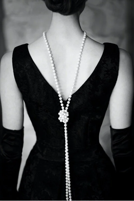 A woman wearing a black sleeveless dress, with a pearl necklace and long black gloves.