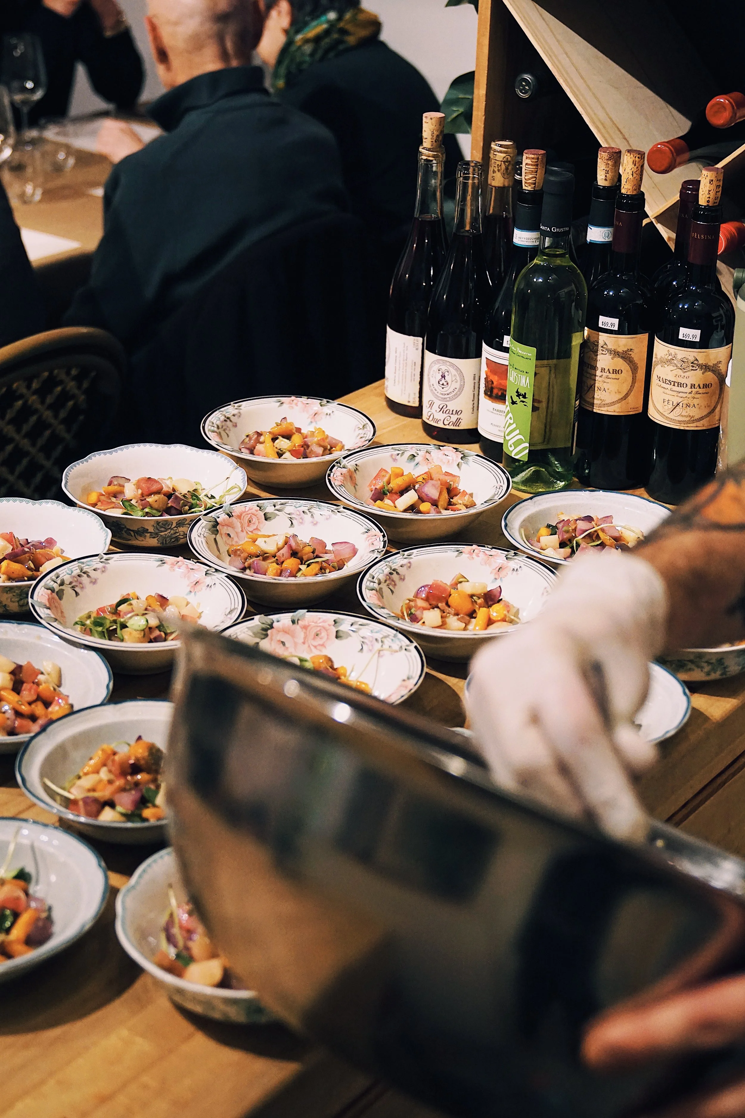 A person serving a colorful vegetable and meat salad into small bowls on a table, with several bottles of wine and glasses in the background.