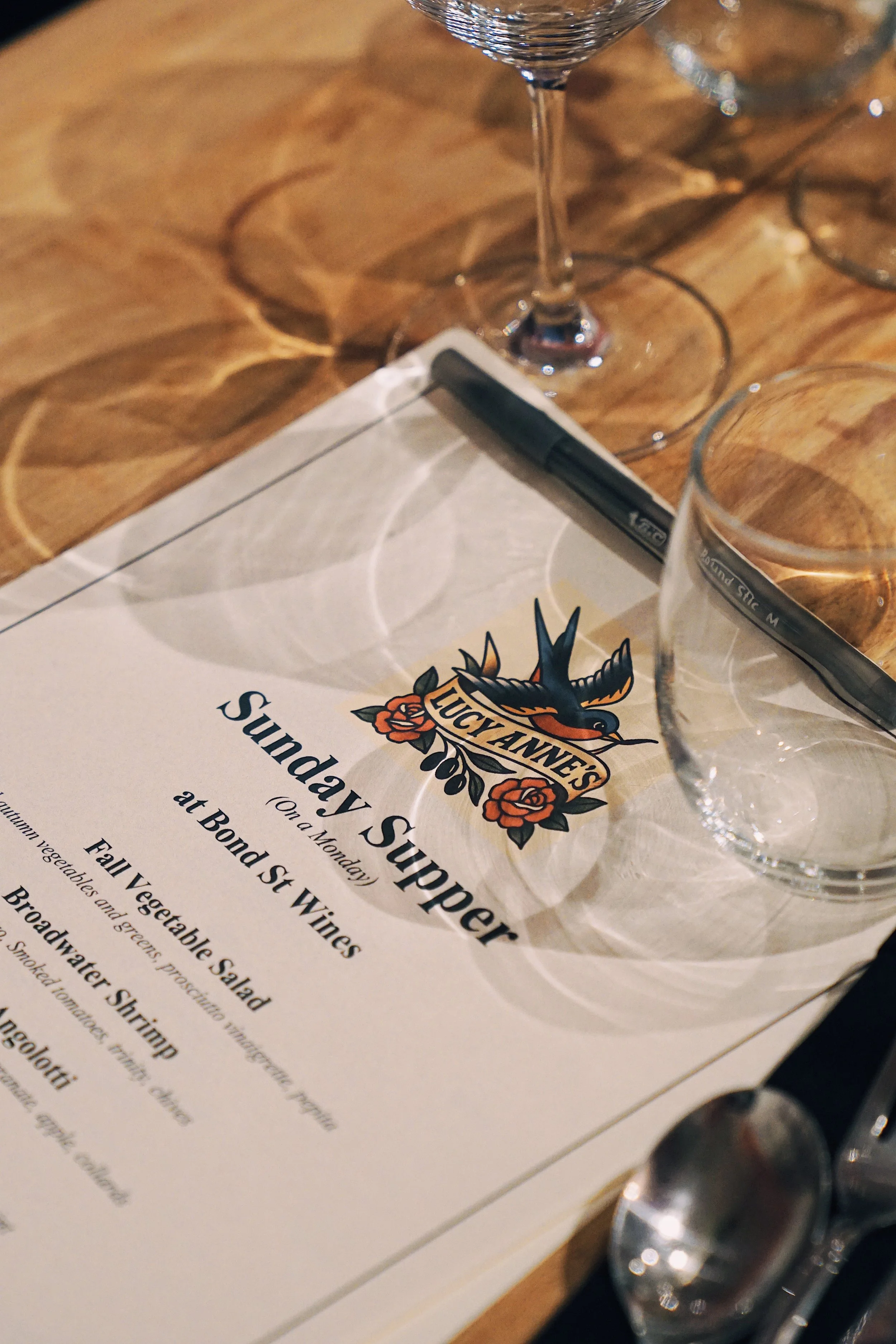 A menu from Lucy Ann's featuring Sunday supper, placed on a wooden table with empty wine glasses and a pen on top.