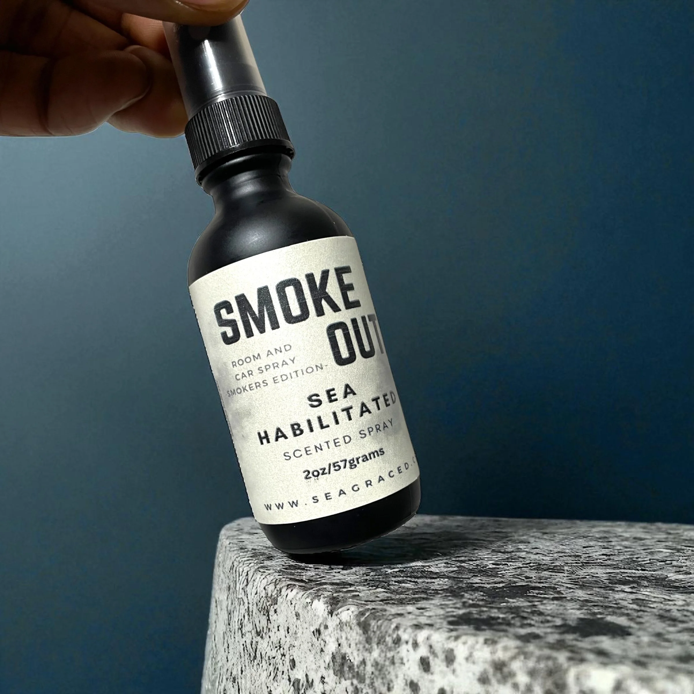 A small black spray bottle with a white label and black cap resting on a weathered wooden surface. The label reads 'SMOKE' in large bold letters, followed by additional text about its use and contents. The background is a plain dark surface.