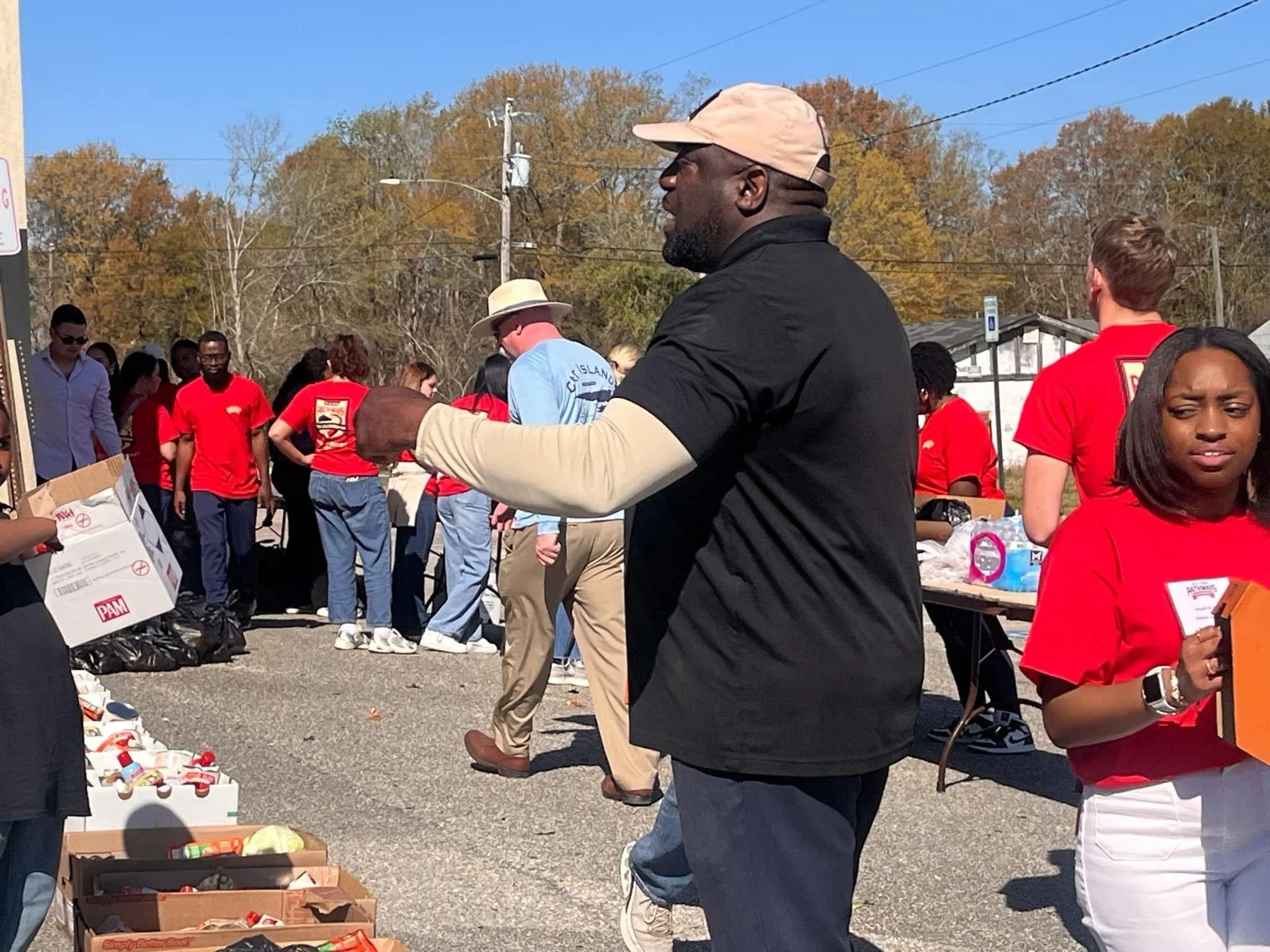 Jeffrey Hulum III directing a community food drive event outdoors; large group of people; some are wearing red shirts with a logo, with tables of supplies and snacks, and trees with fall foliage in the background.