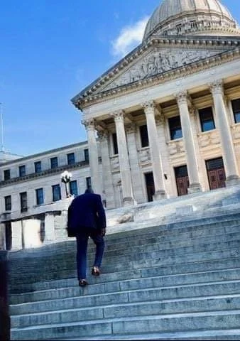 Jeffrey Hulum III in a dark suit climbing the steps at the Mississippi State Capitol after being elected to the State House in 2022. Walking toward a large, historic government building with columns and a dome, under a blue sky.