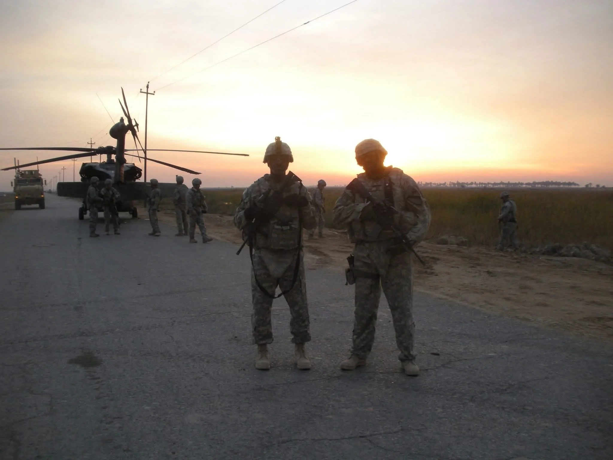 Photo from one of Jeffrey Hulum III's tours of duty with unknown military personnel standing on a road at sunset with a helicopter and others in the background.