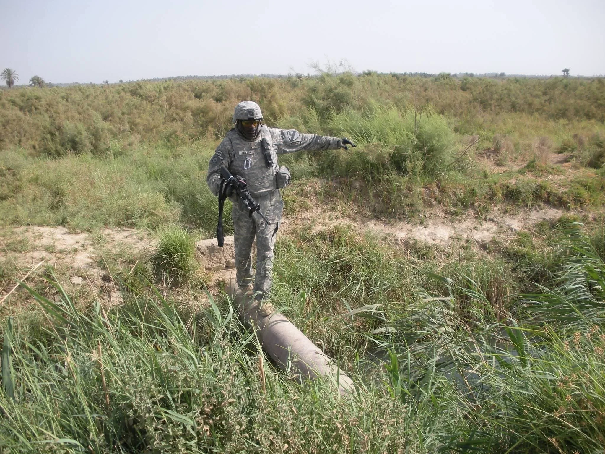 Jeffrey Hulum III in Army training in camouflage uniform holding a rifle, standing on a pipe over a small waterway in a grassy, bushy landscape.