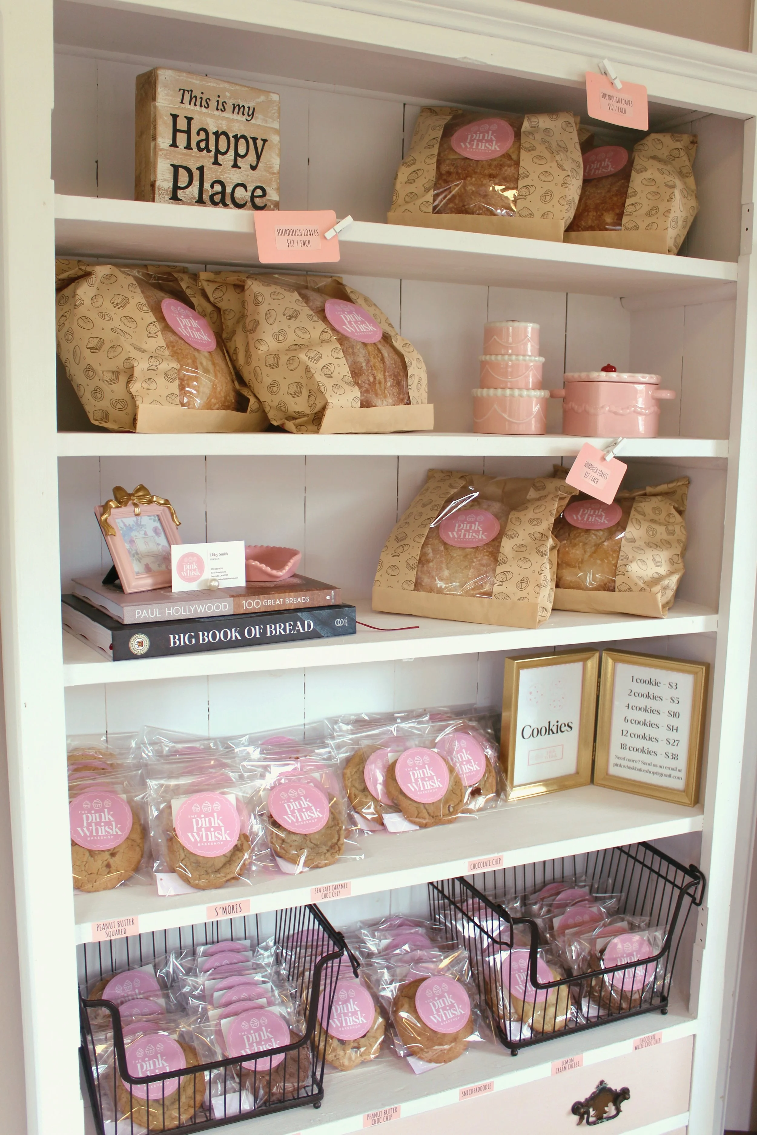 A white shelf filled with pink and brown bakery items, cookbooks, and framed signs, including a sign that reads 'This is my Happy Place.'