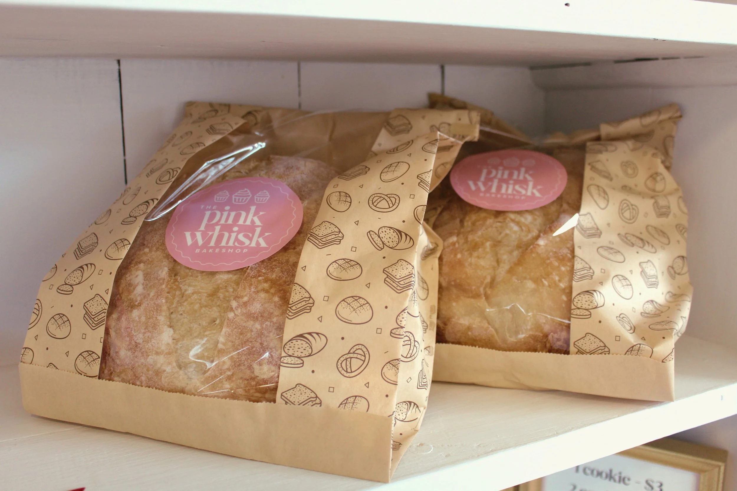 Two loaves of bread inside clear plastic bags with beige and brown patterned paper at the top, labeled with pink stickers that read 'The Pink Whisk Bakeshop,' on a white shelf.