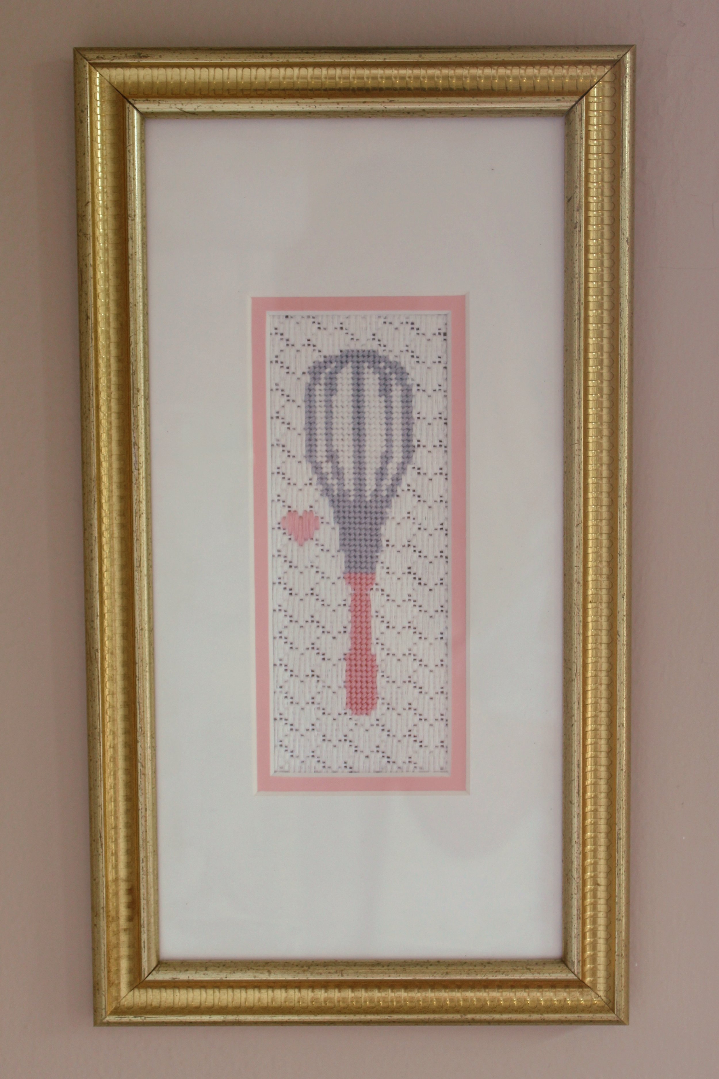 Framed cross-stitch artwork of a hot air balloon with a small pink heart beside it, on beige wall.
