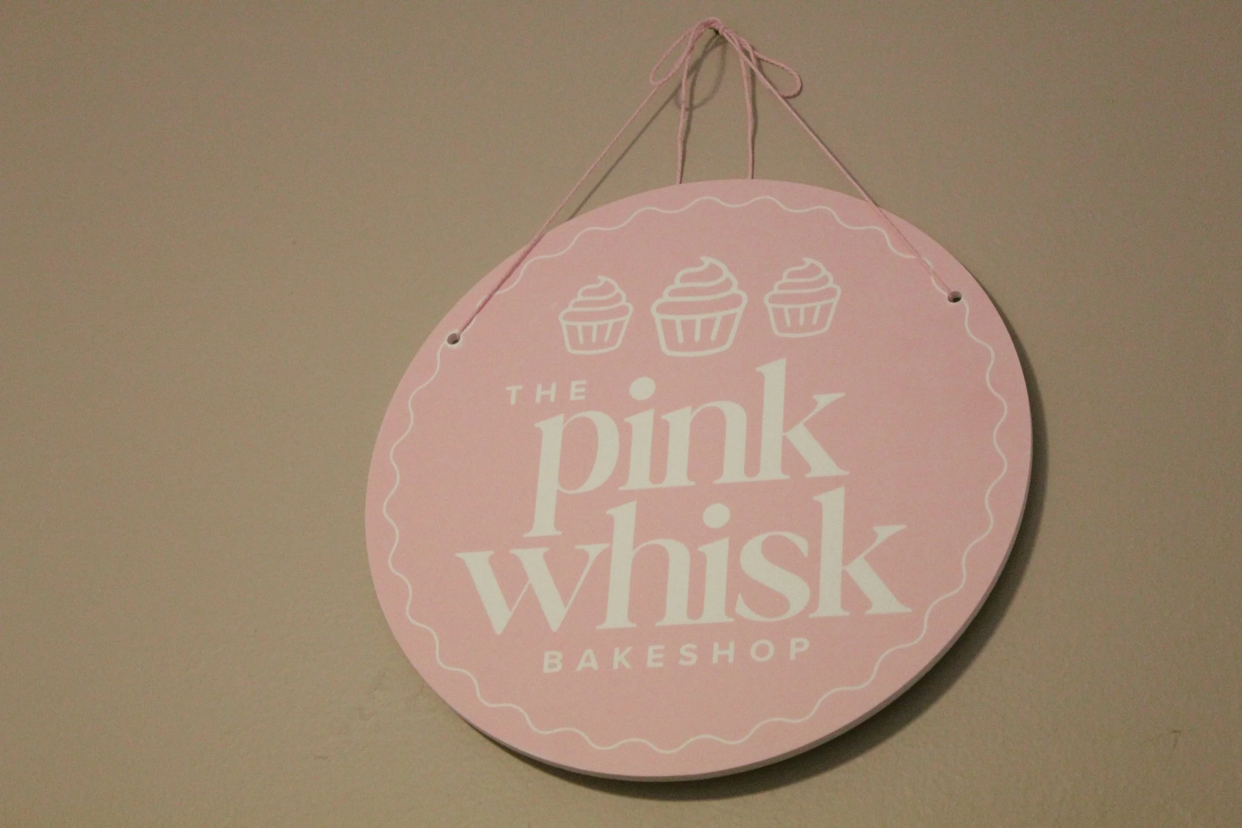 Round pink sign with white text that reads "The pink whisk BAKESHOP" and features three cupcake illustrations at the top, hanging on a beige wall.