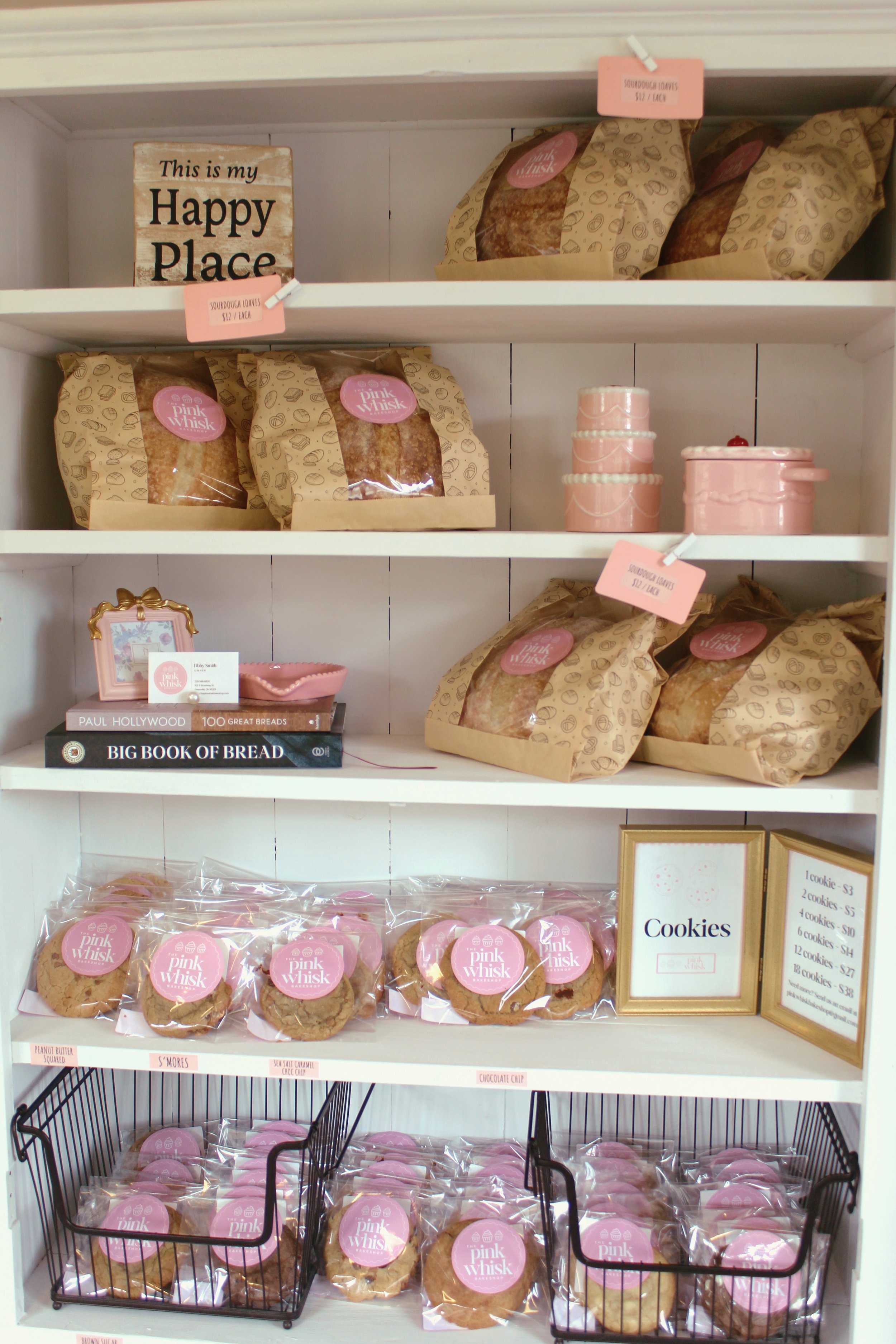 White shelf with pink and brown bakery packages, cookies in clear bags with pink labels, framed cookies sign, and decorative pink containers.
