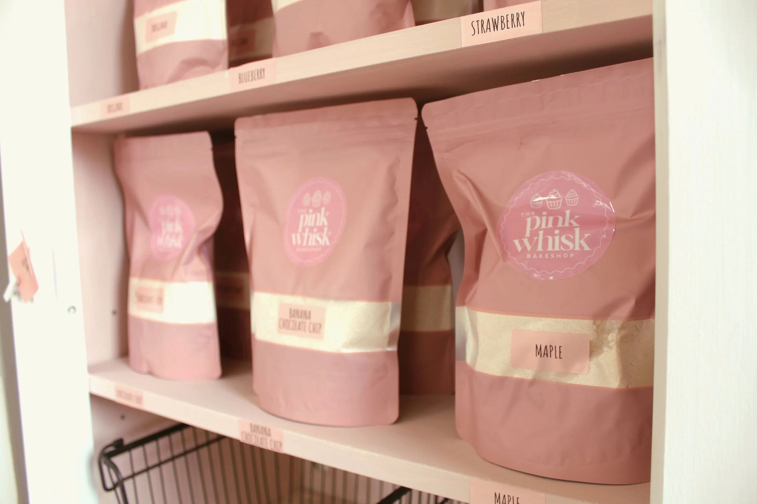 Pink resealable bags of baking mixes on a shelf, labeled with flavors like strawberry, blueberry, banana chocolate chip, and maple from The Pink Whisk Bakeshop.