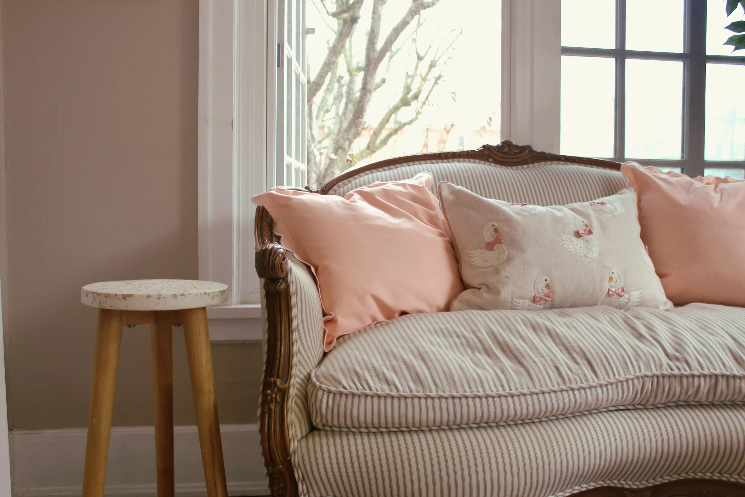A vintage-style sofa with pink and beige pillows, including a decorative pillow with duck embroidery, placed near large windows with a view of trees outside.