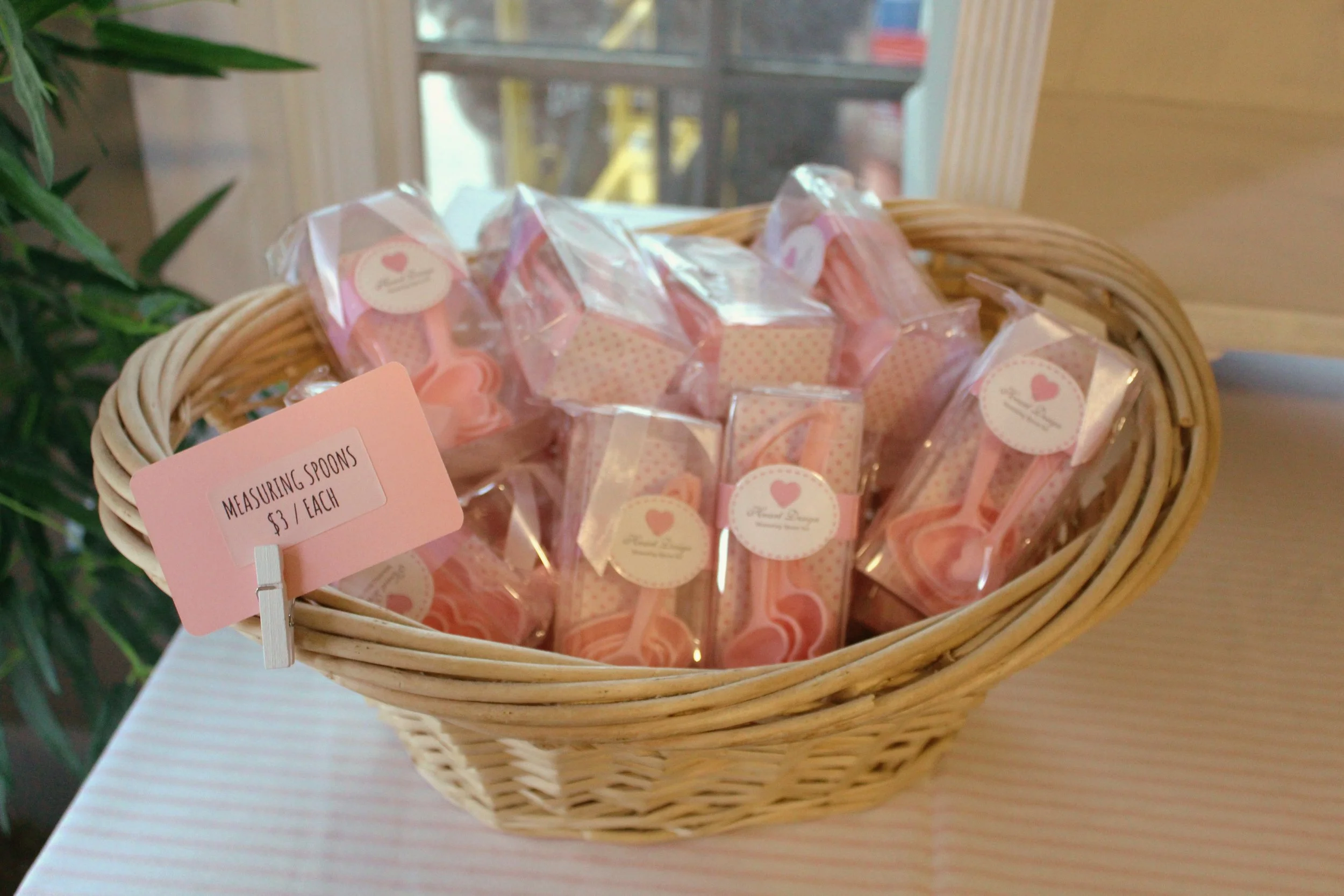 A wicker basket filled with pink and white plastic wrapped heart-shaped measuring spoons, with a pink sign indicating they are $3 each, placed on a pink and white striped table near a window and green plant.