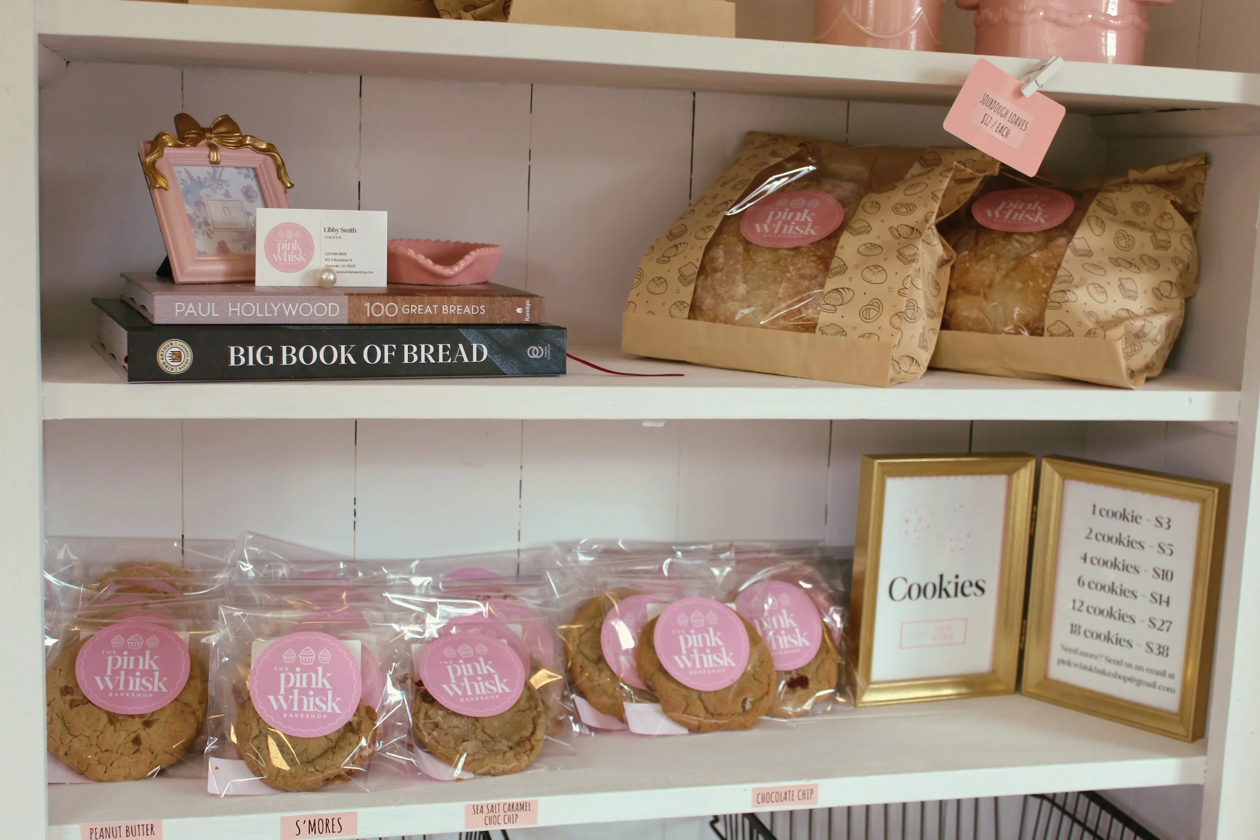 Shelf with baked goods and cookbooks. Top shelf has two loaves of bread in paper bags and a small pink picture frame. Middle shelf has packaged cookies labeled Pink Whisk. Sign with cookie prices on the right. Books titled 'Paul Hollywood 100 Great B