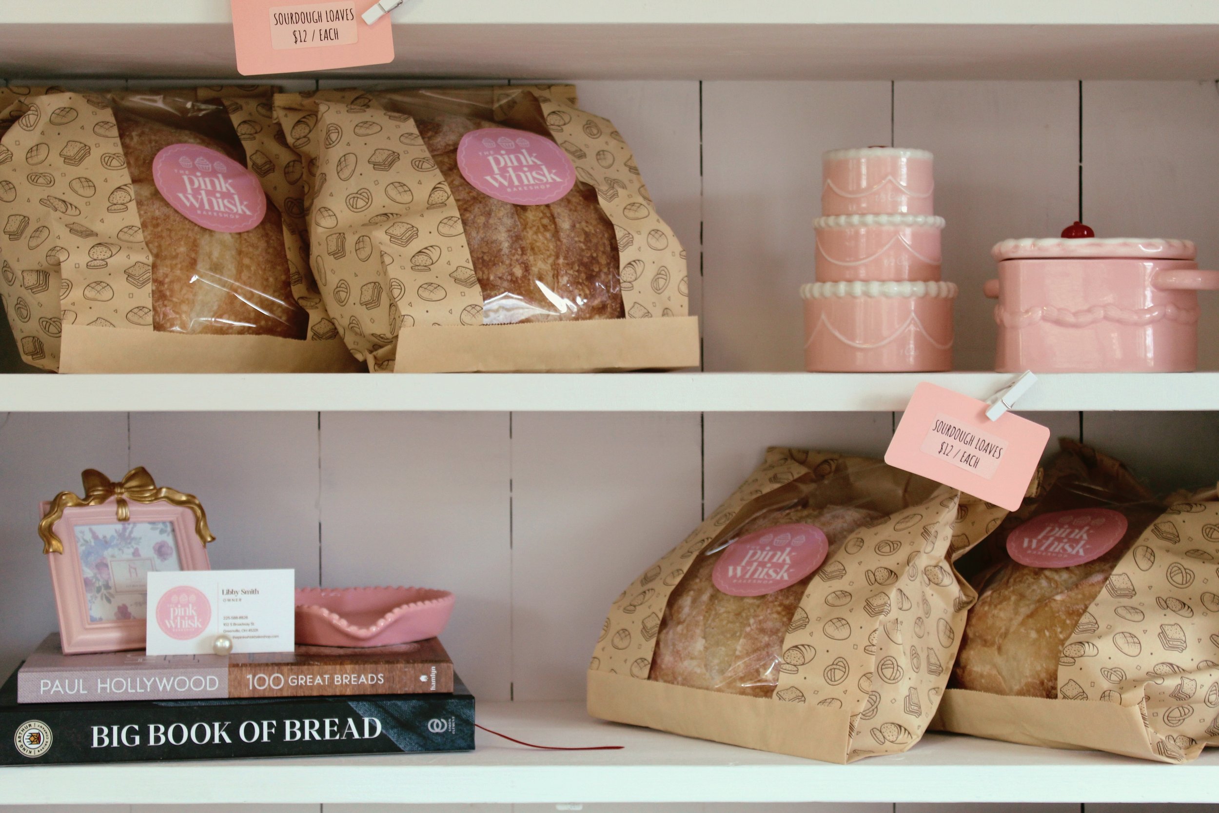 Shelf displaying bags of sourdough loaves with pink labels, pink cake tins and a pink container, a small pink picture frame, a pink bowl, and two large bread books on a white shelf.