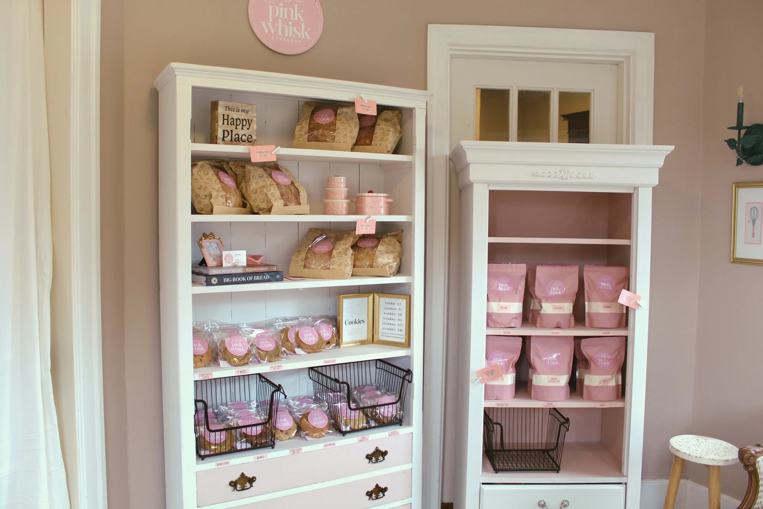 Pastel pink and white shelves displaying packaged baked goods and cookies inside a pink bakery boutique. There are pink labels and a framed price list on the display.