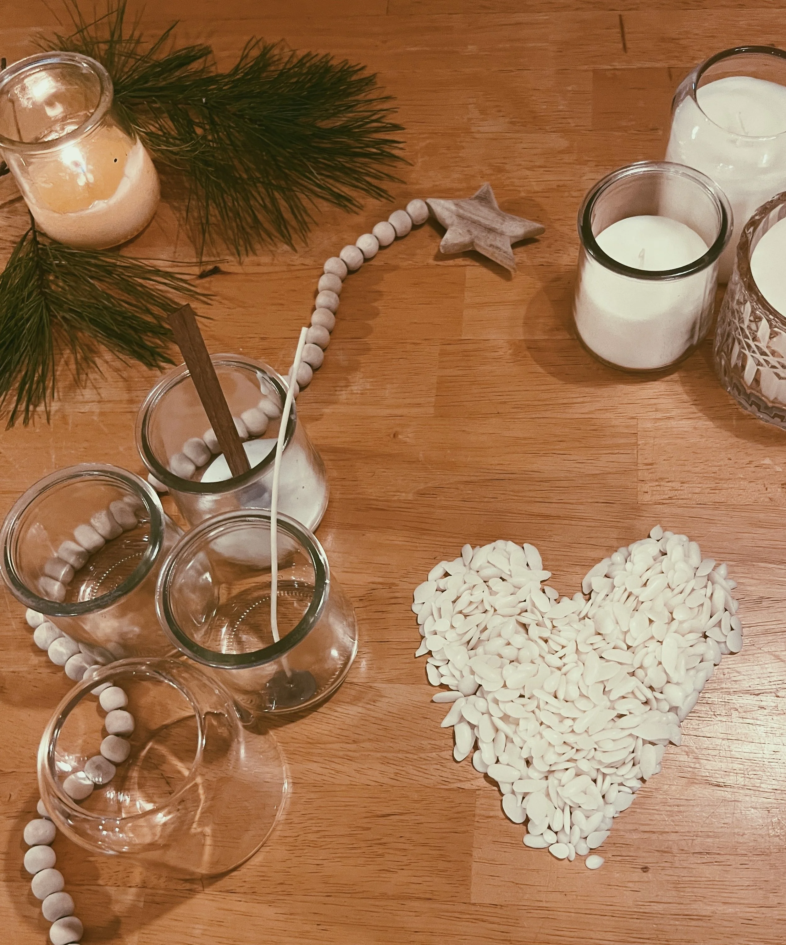 Decorative holiday table setting with white candles, green pine branches, gray star ornament, white beads, and heart-shaped arrangement of white almonds or candies.