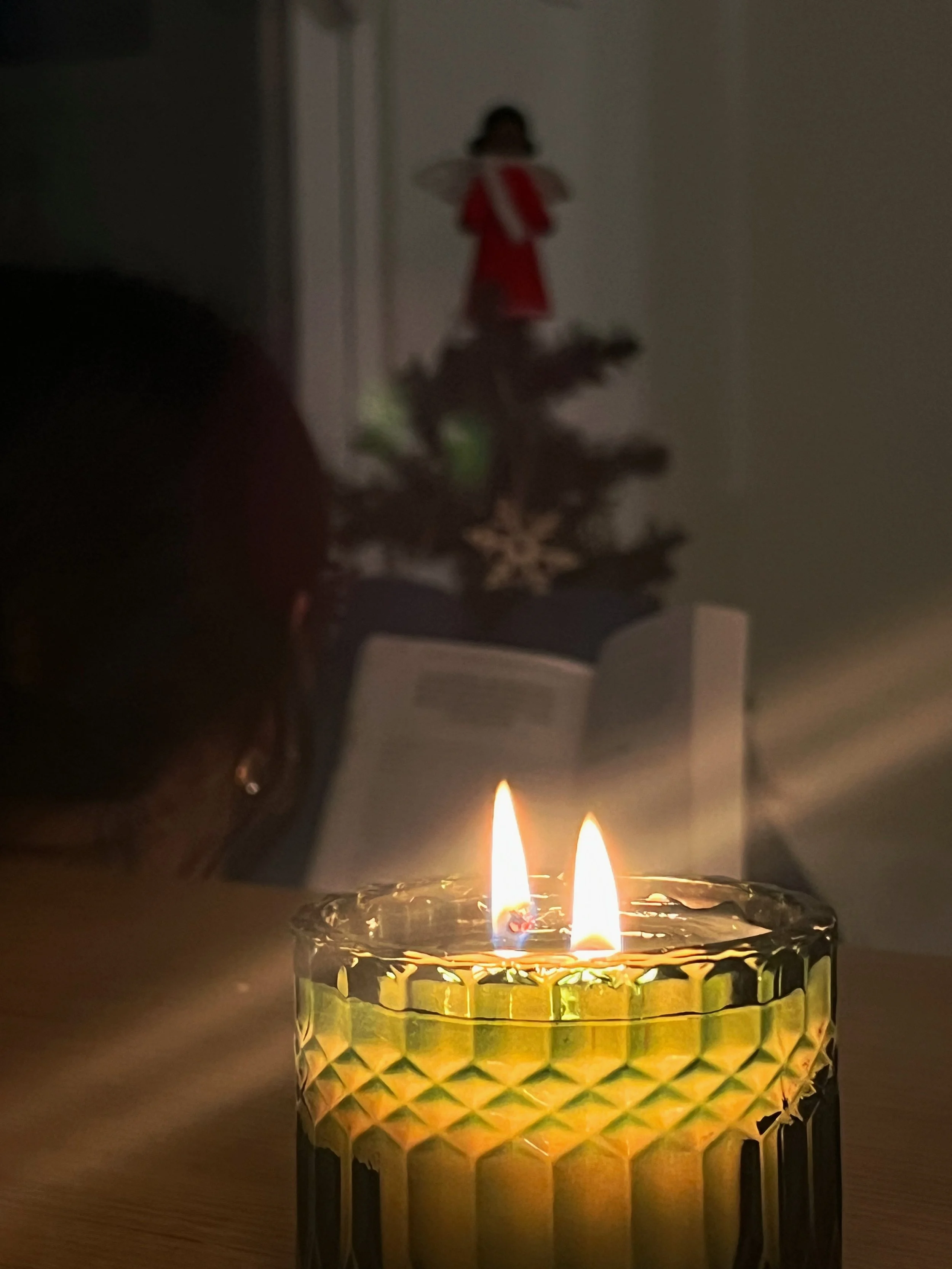 A lit candle in a green, textured glass holder on a table with a small Christmas tree and a blurry figure in the background.