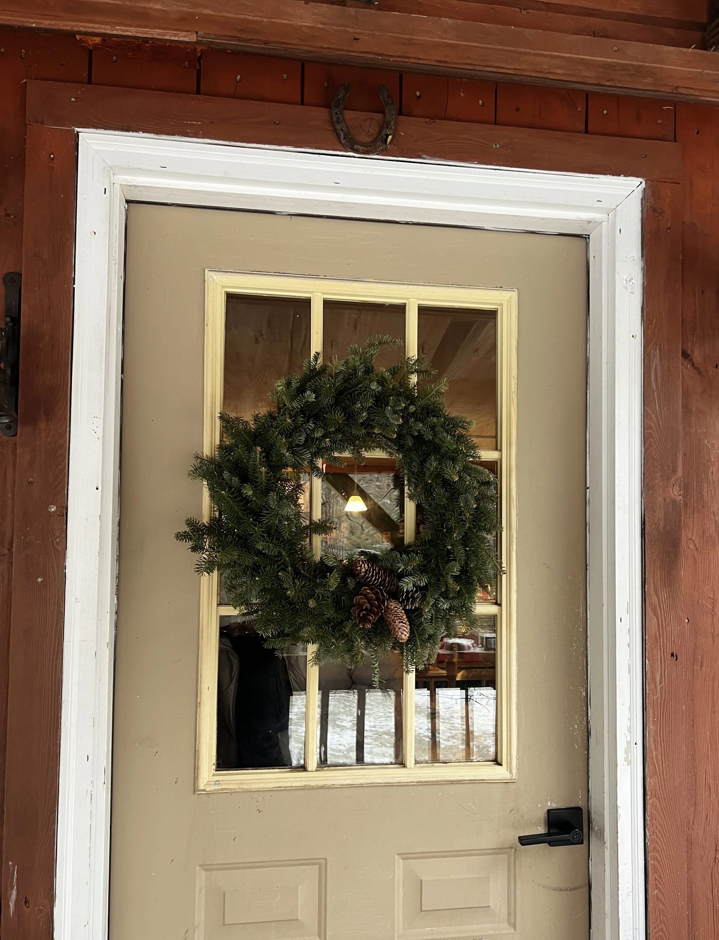 A beige front door decorated with a green Christmas wreath with pinecones, mounted on a red wooden wall with a horseshoe above.