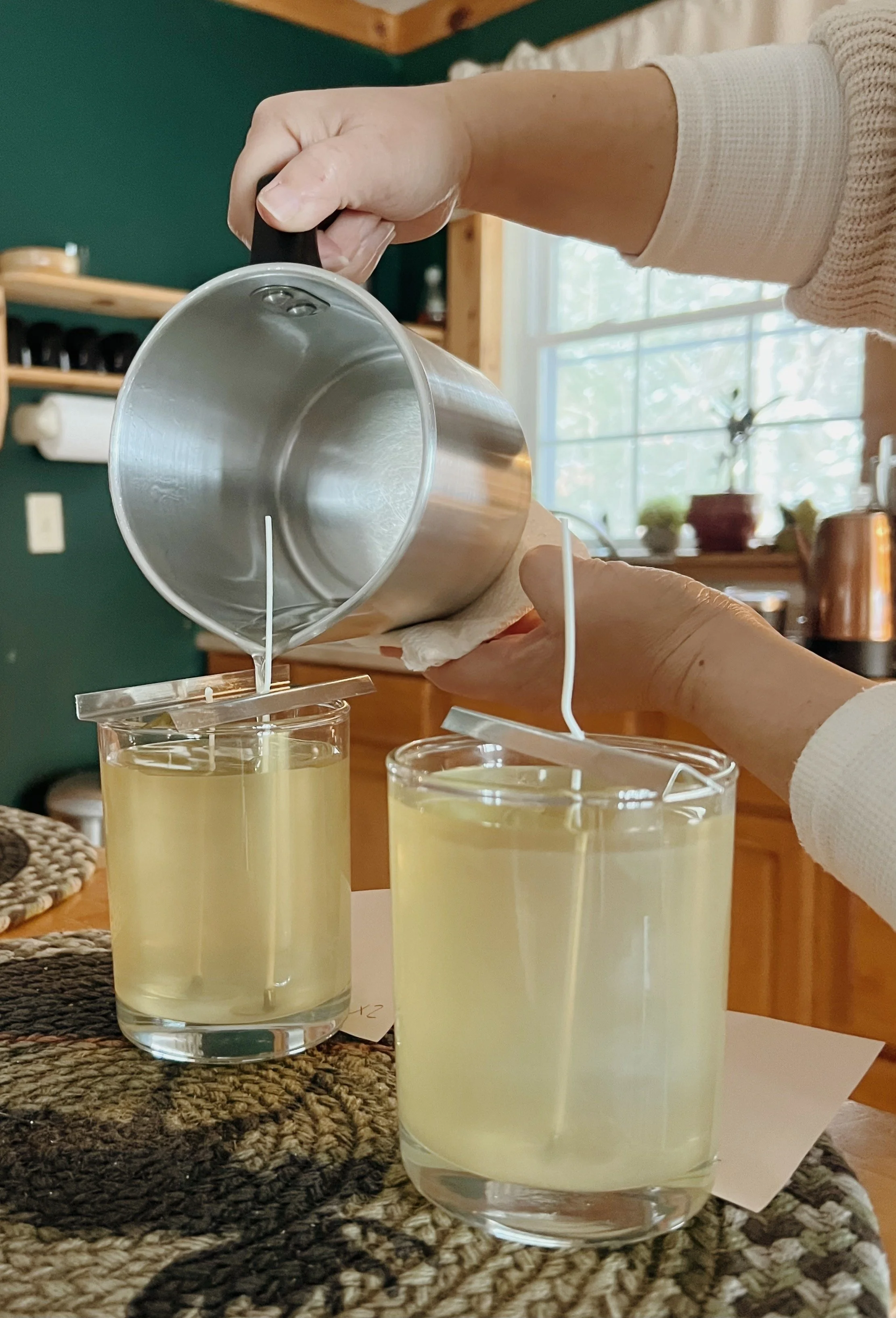 Person pouring a thick, yellow liquid from a metal can into two glass cups, using a small squeegee to hold the paper towel around the can's spout. The setting appears to be a cozy kitchen with natural light coming through a large window.