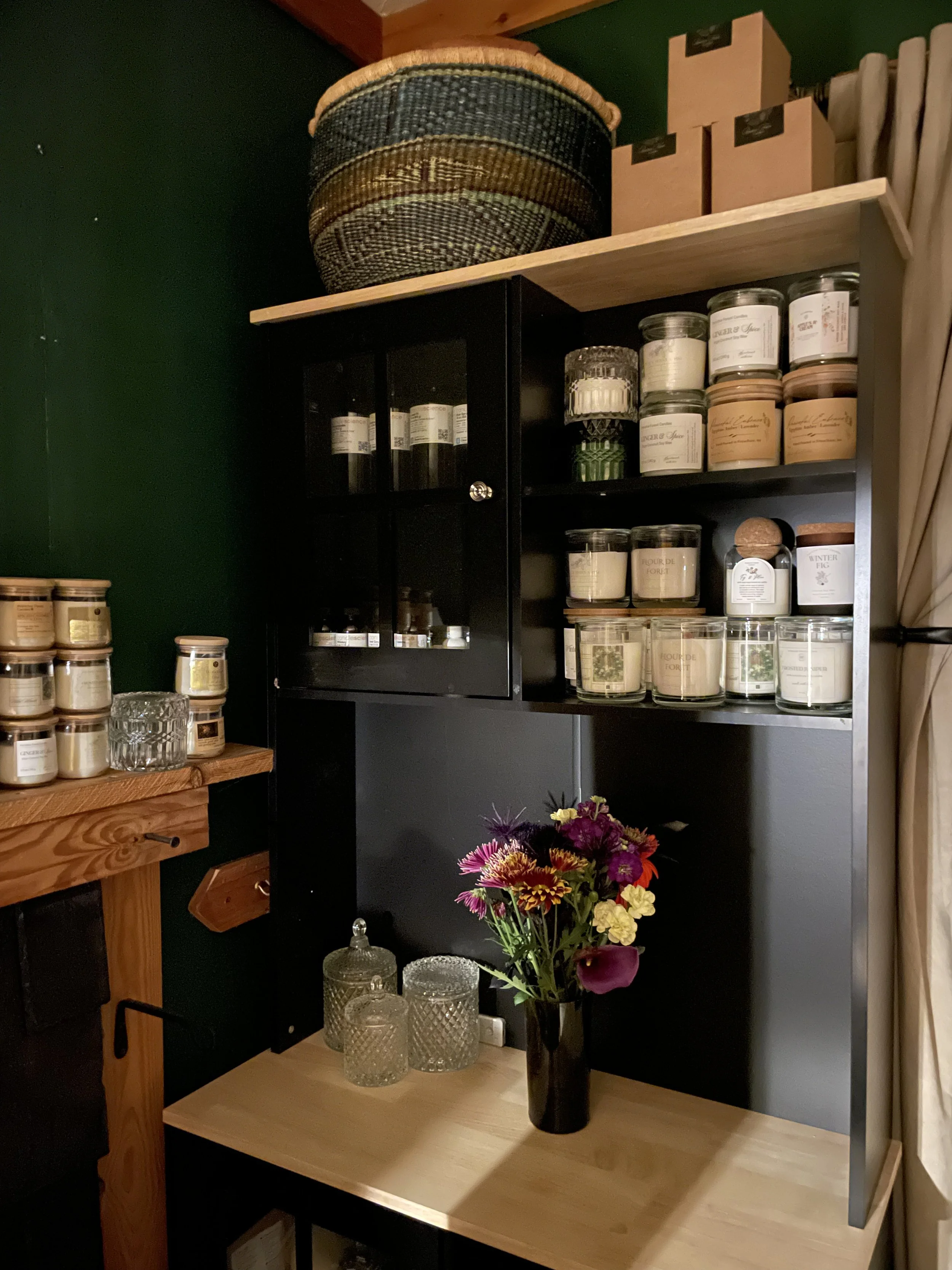 A corner shelf unit with jars of spices, candles, and other items, alongside a wooden table holding a vase of colorful flowers and glass jars.