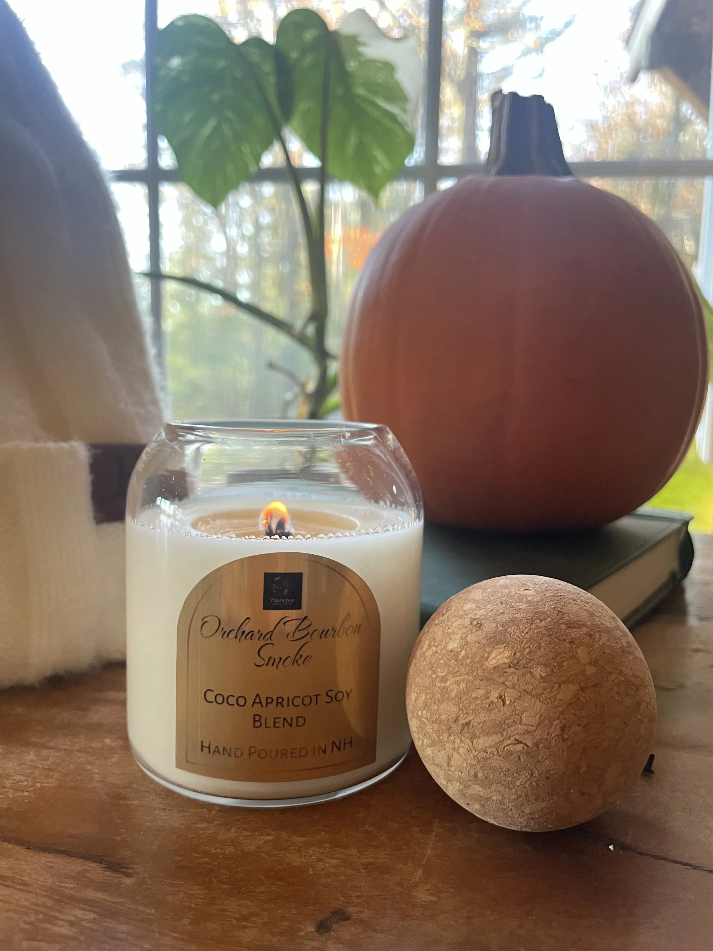 A lit candle labeled 'Orchard Bourbon Smoke Coco Apricot Soy Blend Hand Poured in NH' placed on a wooden surface. Behind it, a round pumpkin, a small decorative ball, a book, and a green plant with large leaves are visible. A window with an autumn la