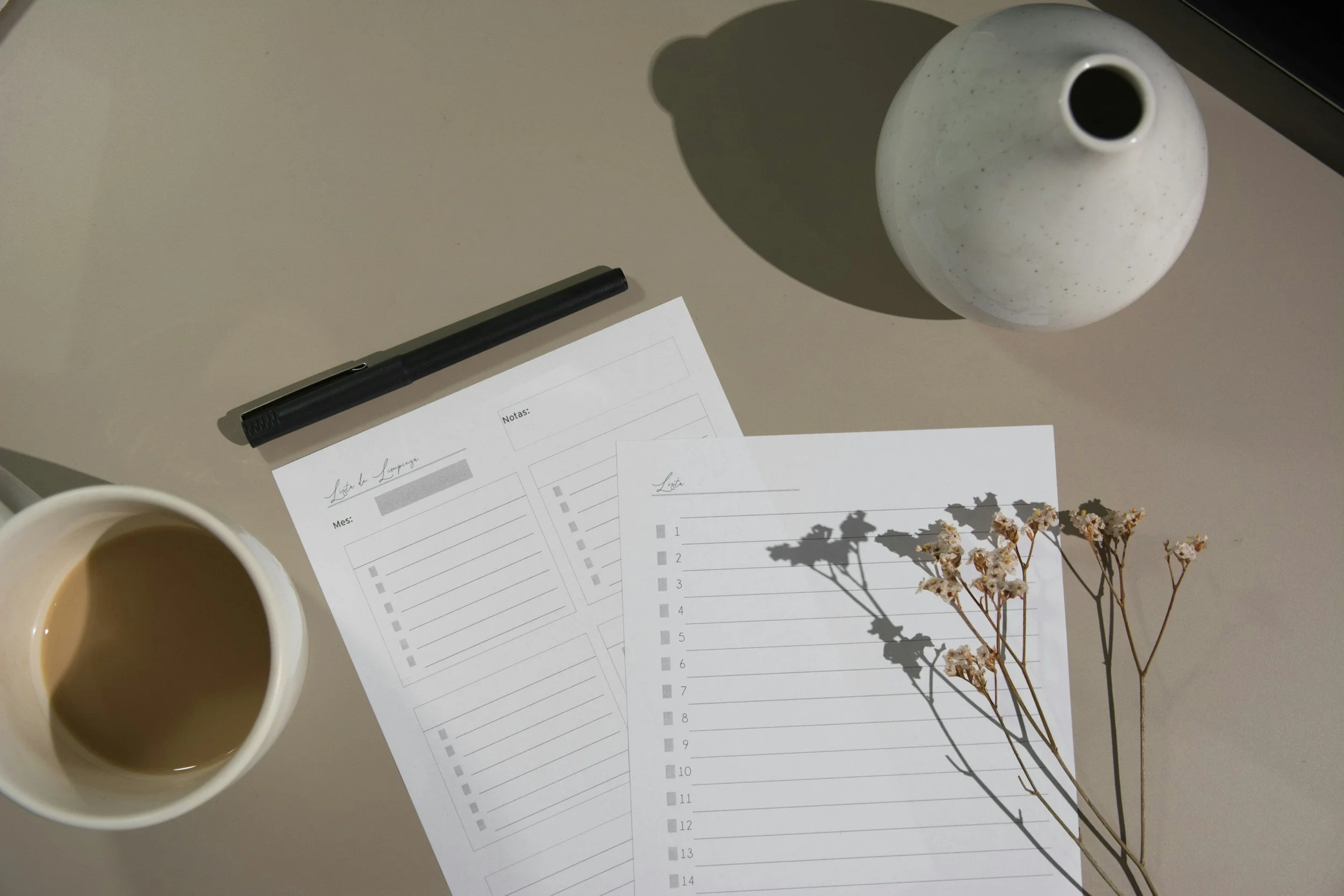 Top view of a desk with a black pen, two sheets of paper with checklists, a cup of coffee, a white vase, and dried flowers casting shadows.