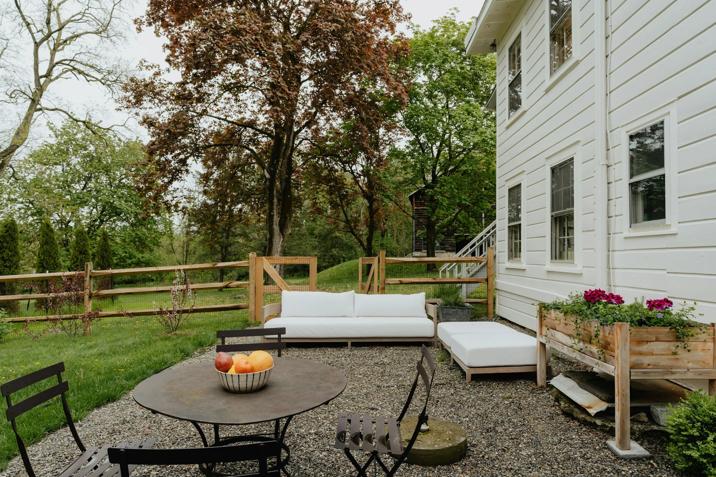 Outdoor backyard patio with round metal table and four chairs, bowl of apples on table, white cushioned sofa and ottoman near white house, surrounded by wooden fence, green trees, and grass