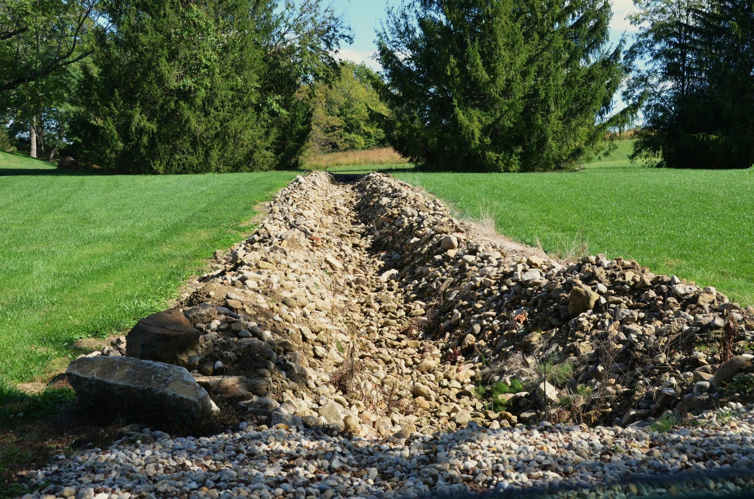A grassy field with a large pile of rocks and gravel in the center, surrounded by trees under a clear blue sky.
