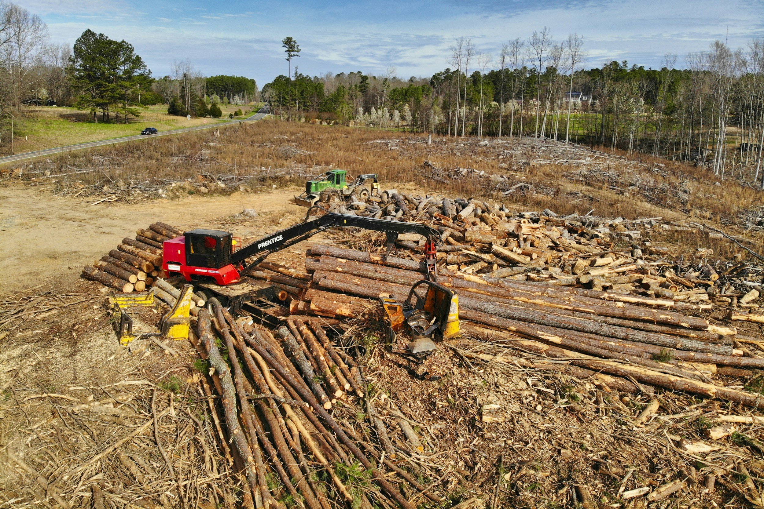 Logging site with a red and black logging machine stacking logs, and a green tractor in the background. Wood logs and debris are scattered across a cleared area surrounded by trees and a road.