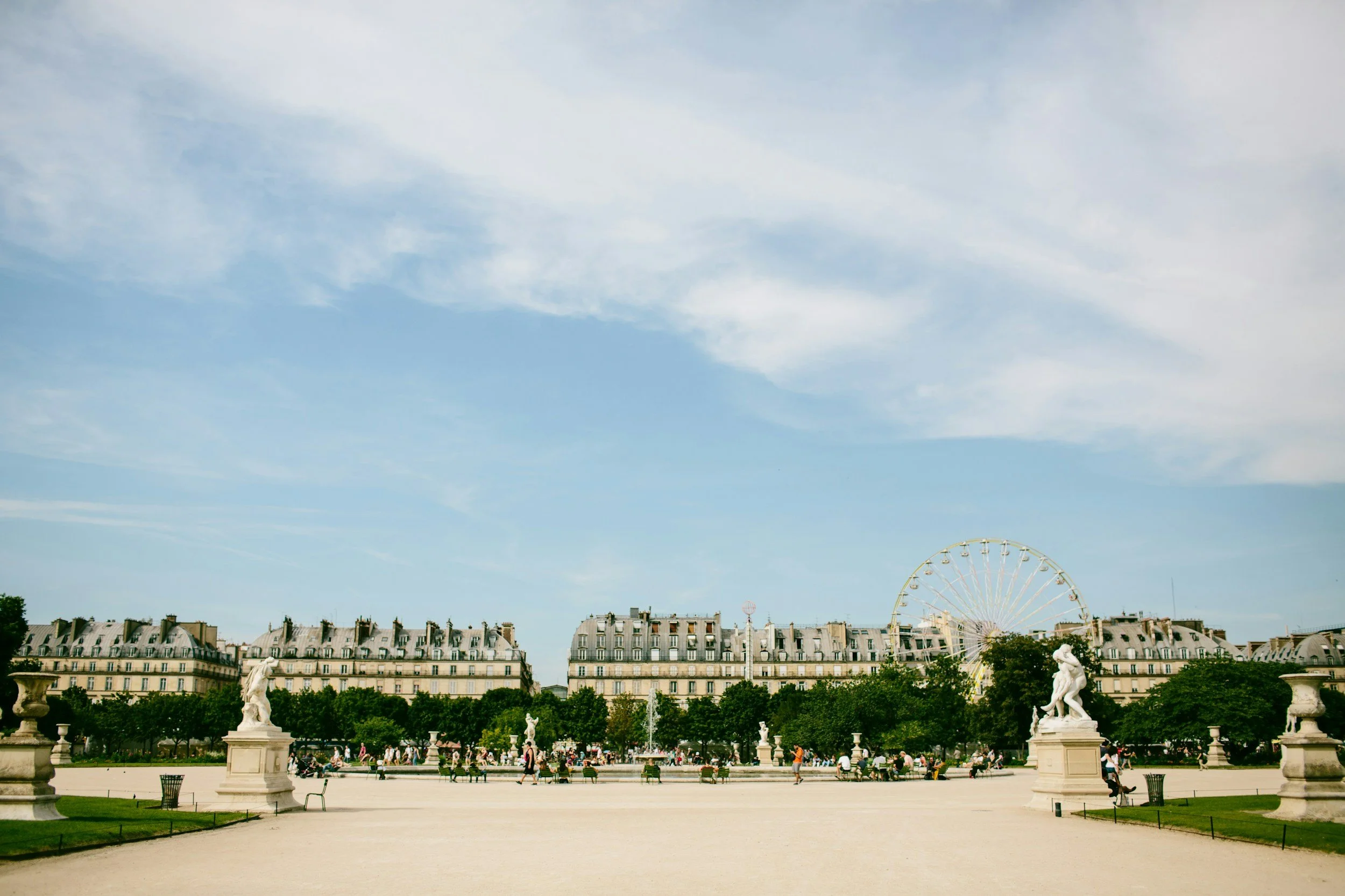 Vue d'une place publique en Paris avec des statues, un grand manège, des arbres et des bâtiments haussmanniens sous un ciel partiellement nuageux.