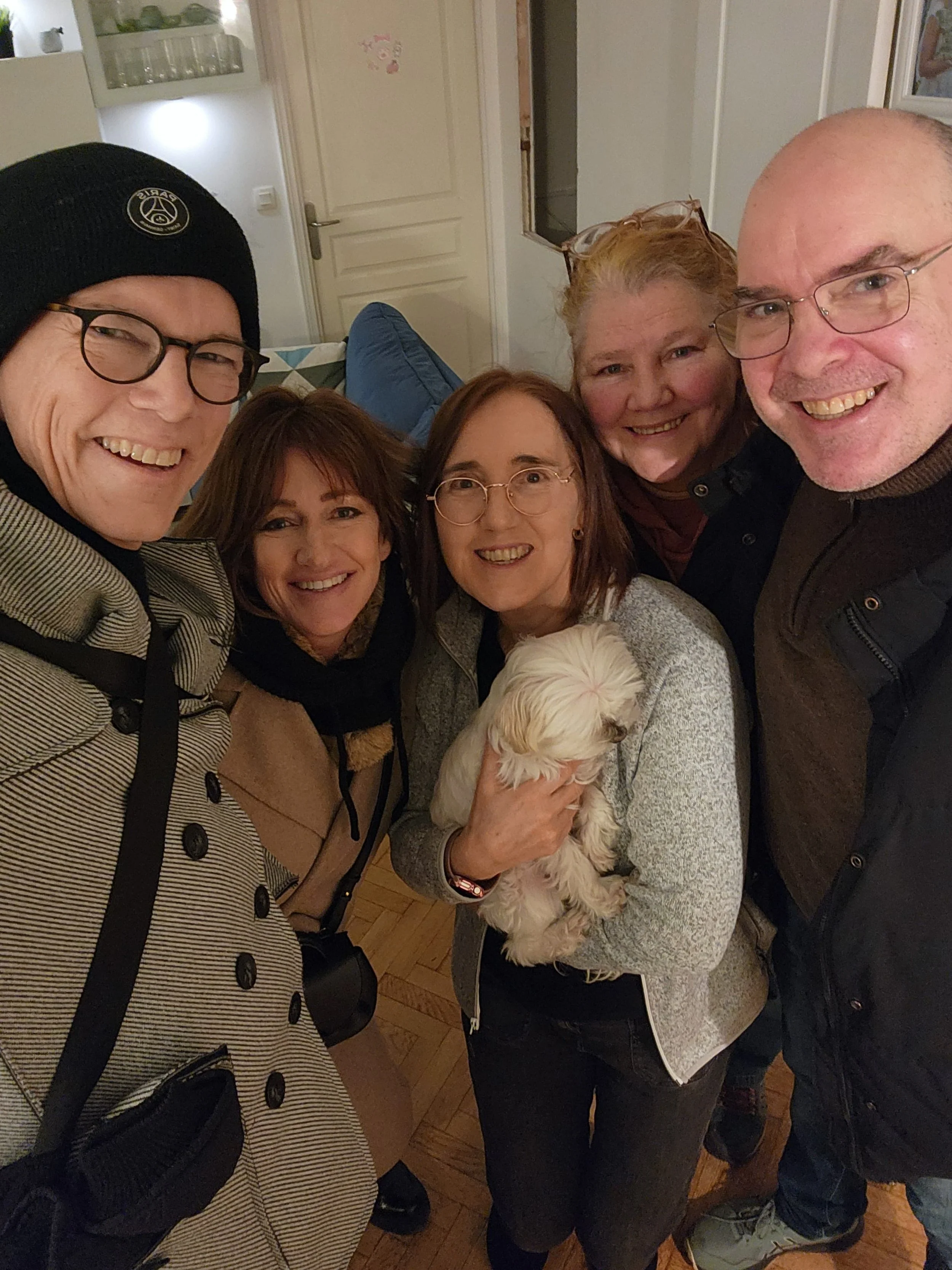 Groupe de cinq personnes souriantes, deux hommes et trois femmes, posant pour une photo en intérieur, avec un chien blanc que l'une des femmes tient dans ses bras.