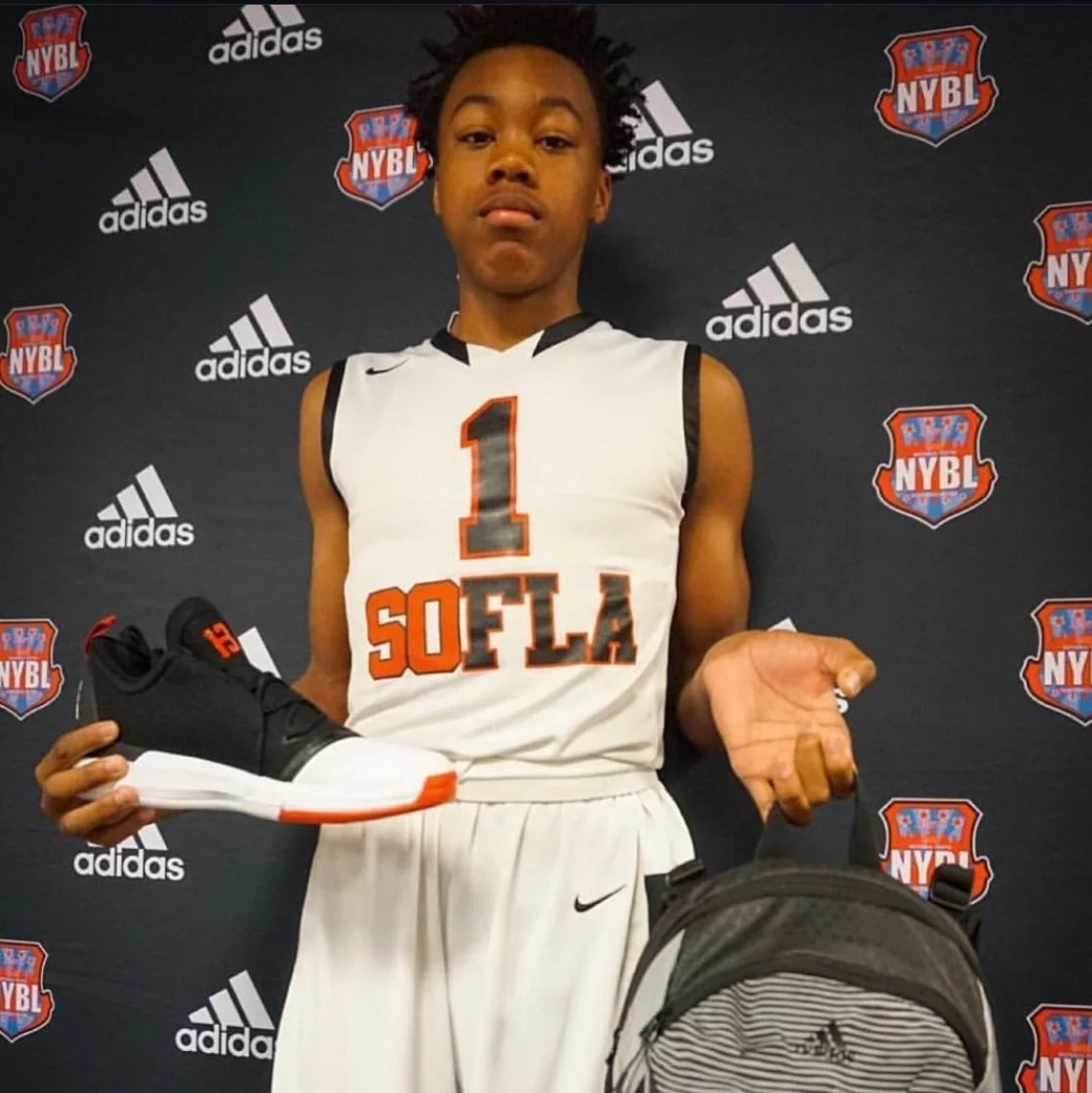 A young basketball player in a white jersey with 'S.O.F.L.A' and the number 1, standing in front of a backdrop with 'NYBL' and 'adidas' logos, holding a black and white sneaker and a backpack.