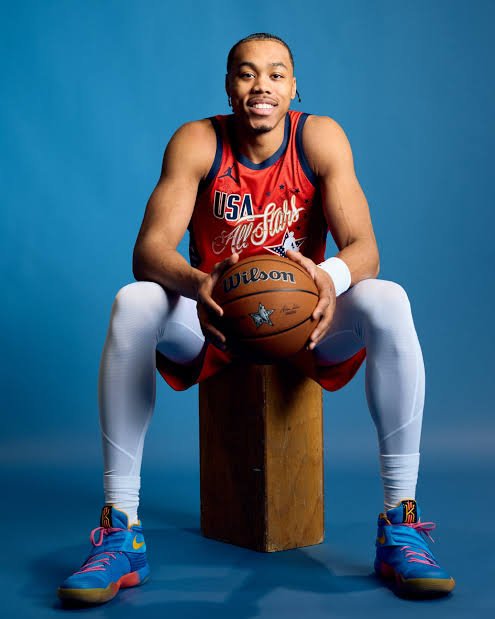 A male basketball player sitting on a wooden block, holding a basketball, wearing a USA All Stars basketball jersey, white tights, and colorful sneakers, against a blue background.