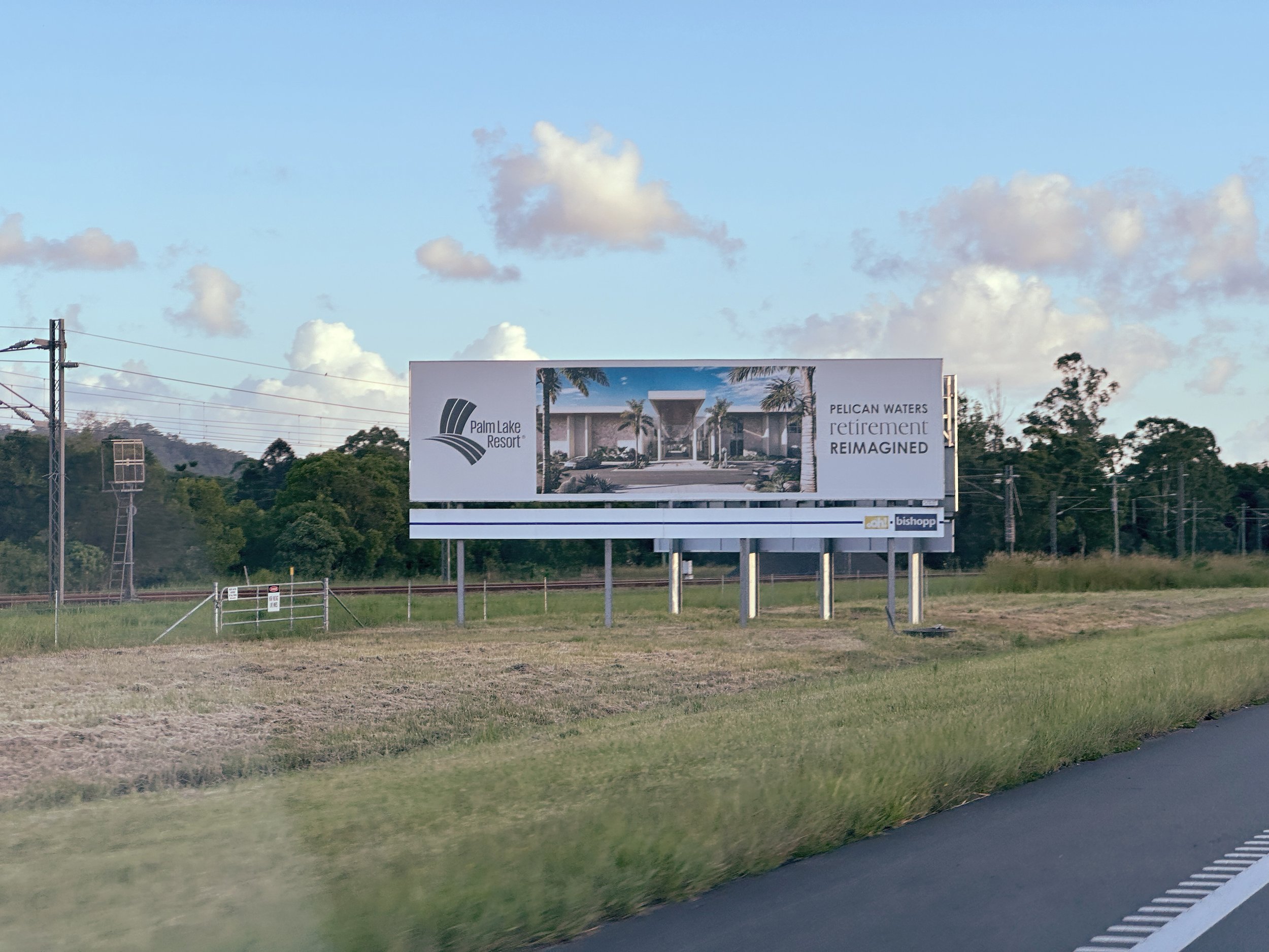 A billboard on the side of a highway advertising Palm Lake Resort, featuring an artist's rendering of a retirement community, with trees and power lines in the background.