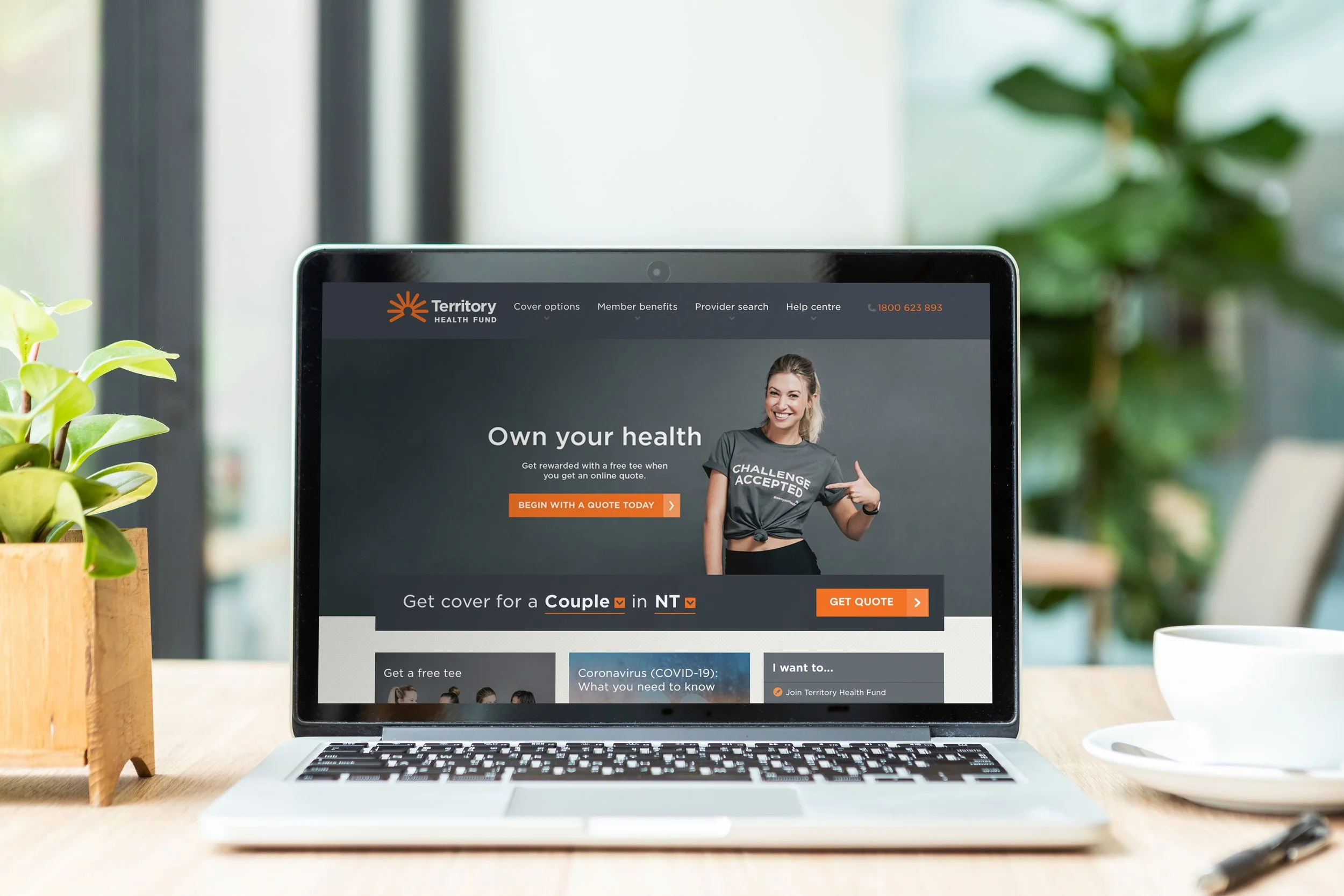 Laptop displaying the Health Fund website on a wooden desk, with a potted plant on the left and a white cup and saucer on the right.