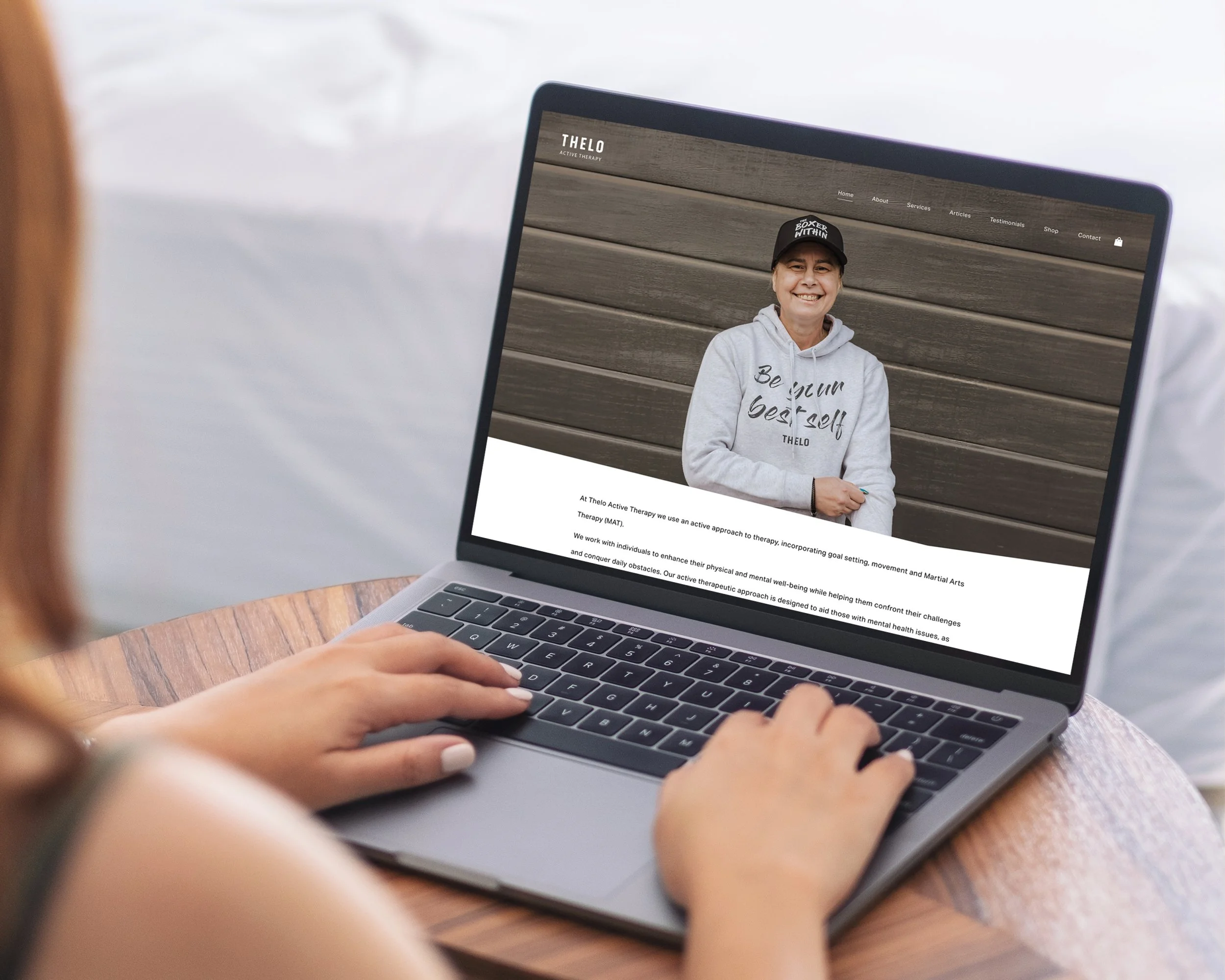 Person using a laptop on a wooden table, viewing a webpage with an image of a woman in a hoodie and cap, smiling against a wooden wall background.