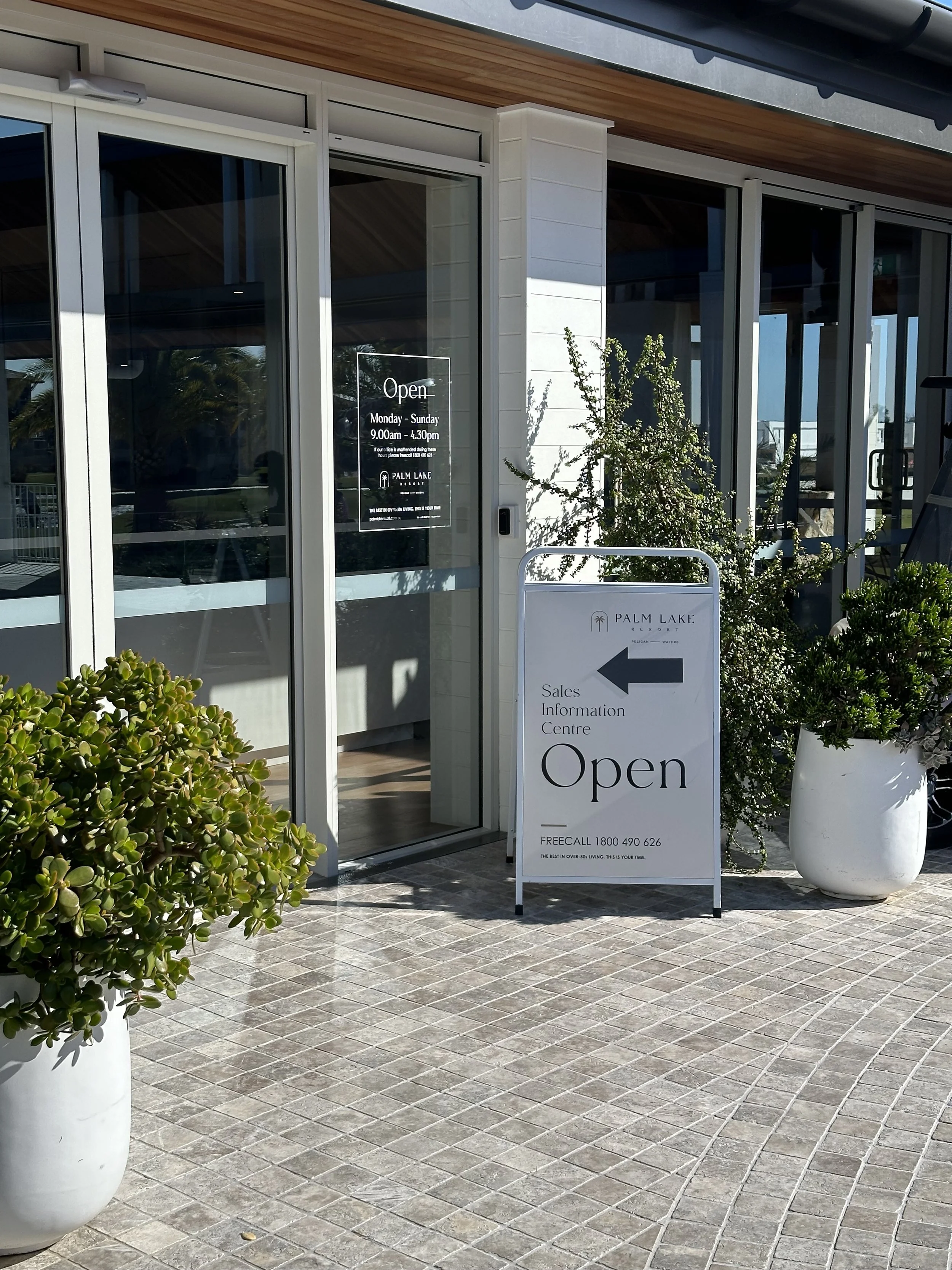 Entrance to a resort sales office with a large white sign indicating the sales information centre is open, and a glass door with a sign showing the resort's hours, surrounded by potted plants and outdoor tiles.