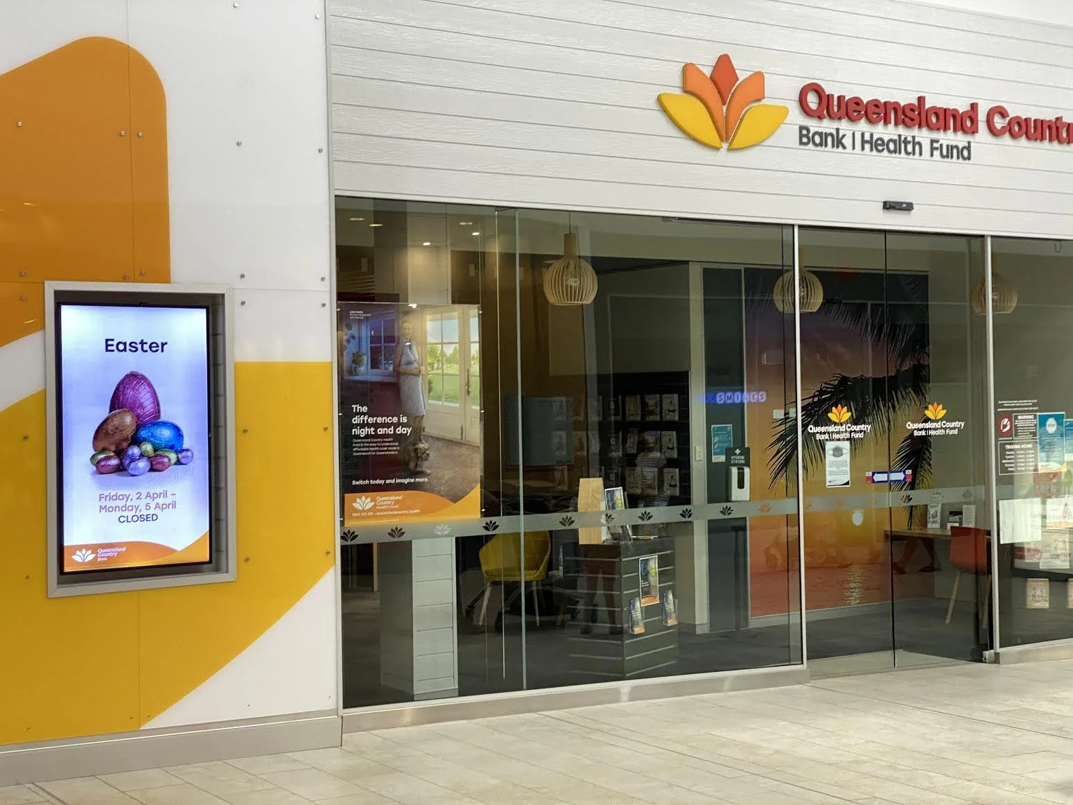 Exterior view of Queensland County Bank and Health Fund office with glass doors, a digital sign displaying Easter holiday hours, and a poster on the window with a palm tree and sunset background.