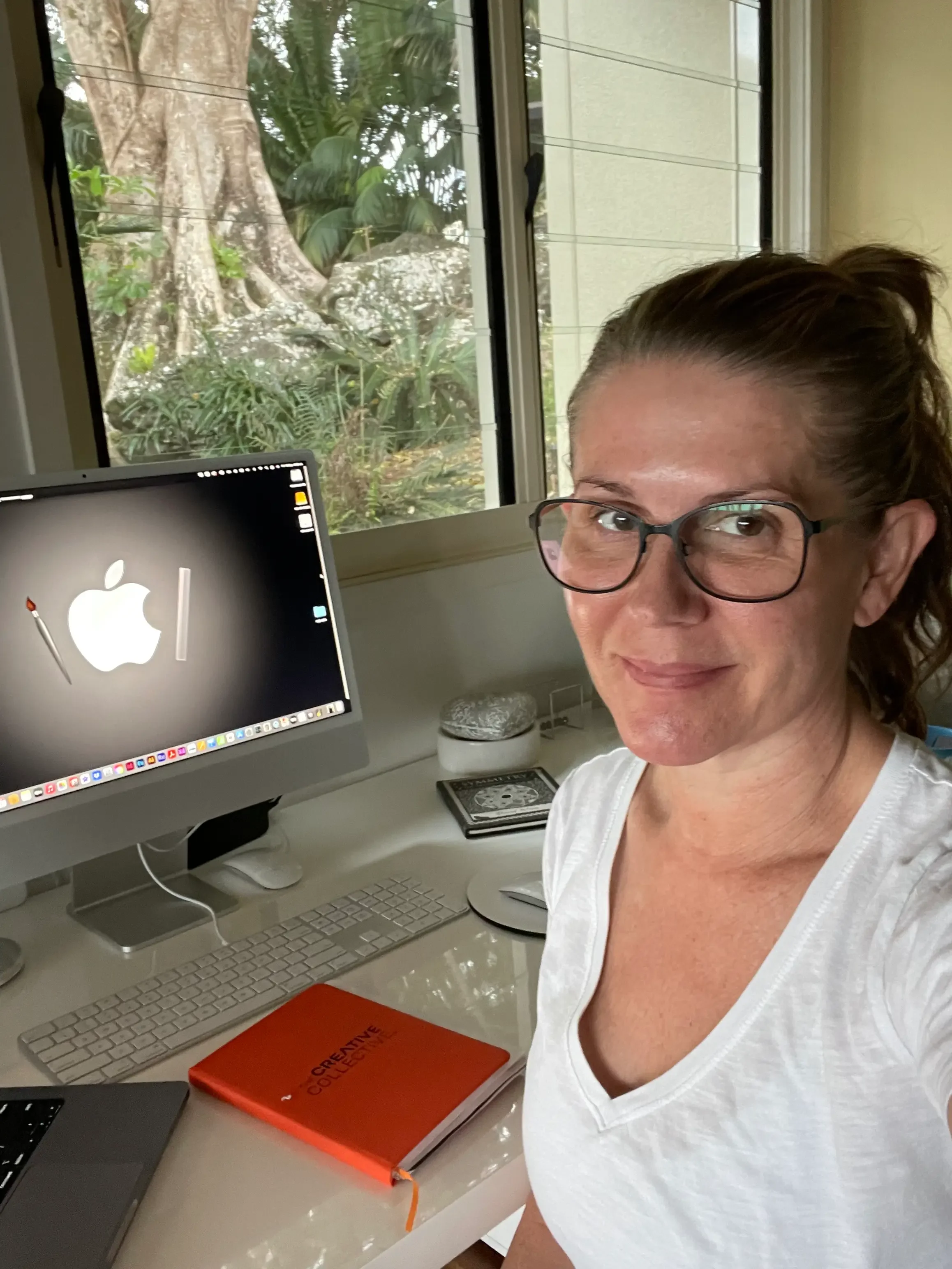 A woman with glasses sitting at a desk in front of an iMac computer with a forest visible outside the window. The desk has a white keyboard, and notebook.