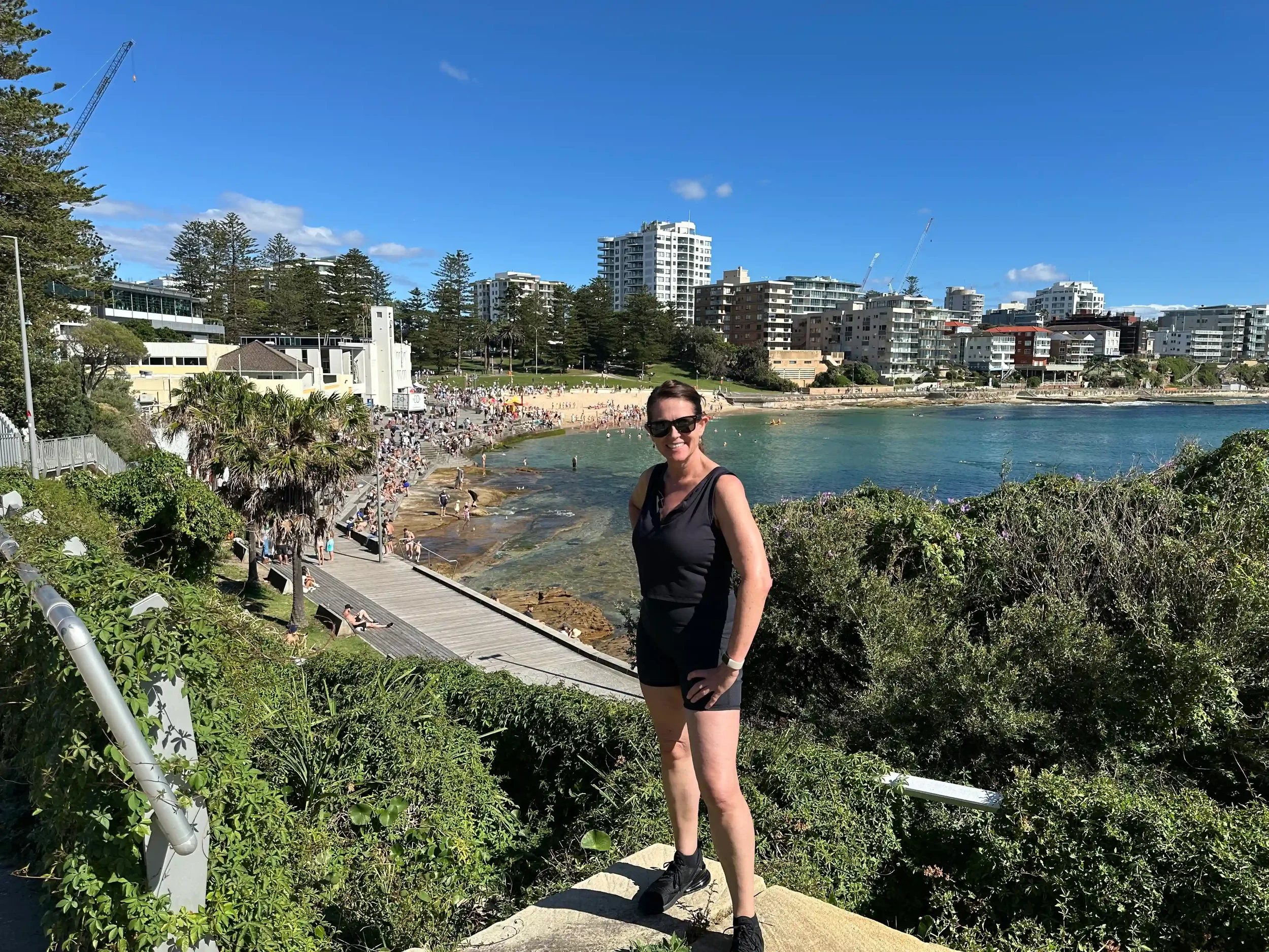 A woman with sunglasses, black shirt, and shorts standing on a coastal trail with a city skyline and beach in the background on a sunny day.