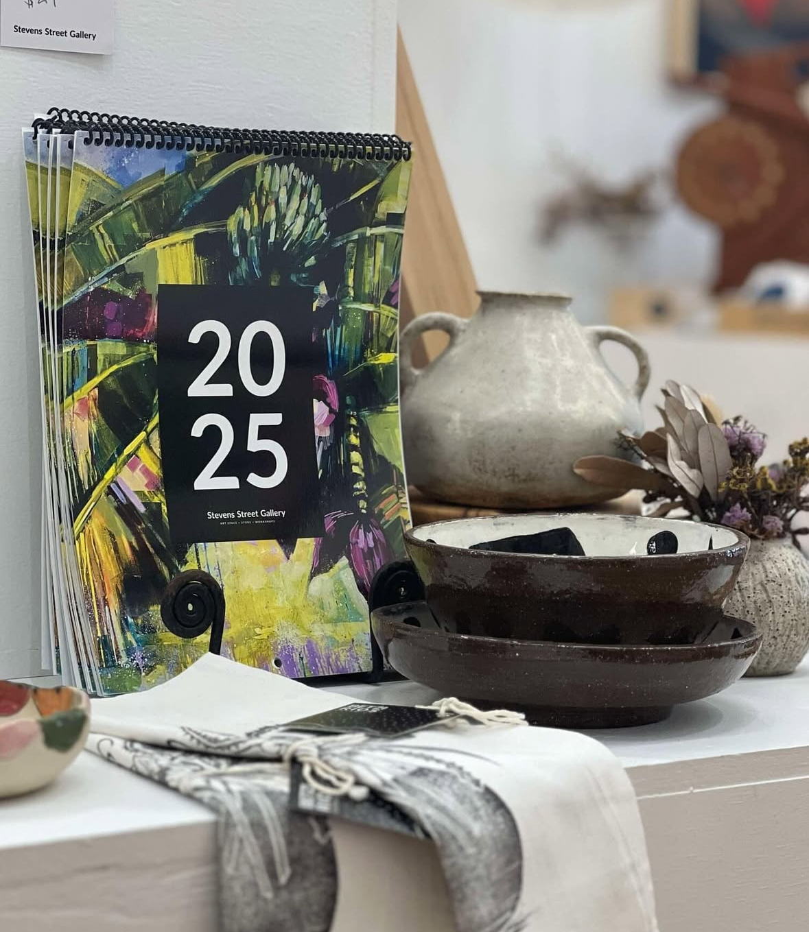 Brochure or catalog titled '2025' from Stevens Street Gallery, displaying colourful abstract artwork, along with ceramic pottery and dried flowers on a table.