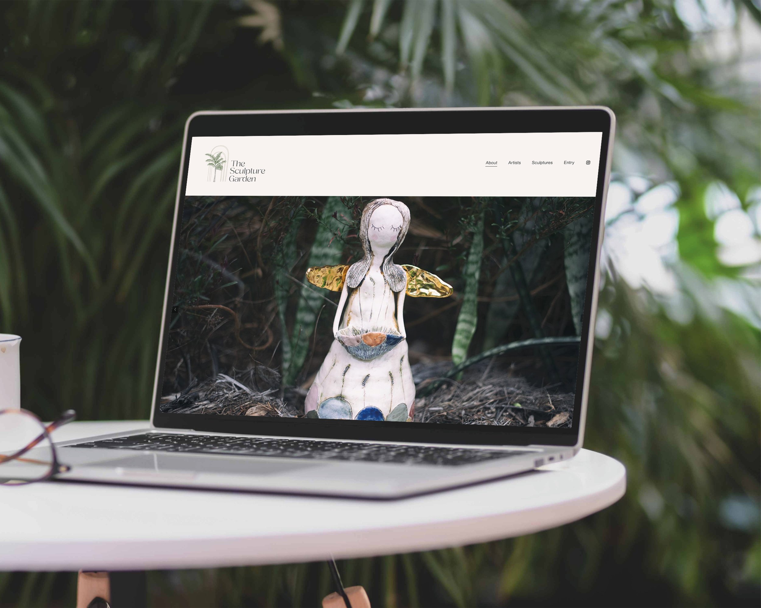 Laptop displaying a website for The Sculpture Garden with an image of a ceramic sculpture of a fairy or angel on a natural outdoor background.
