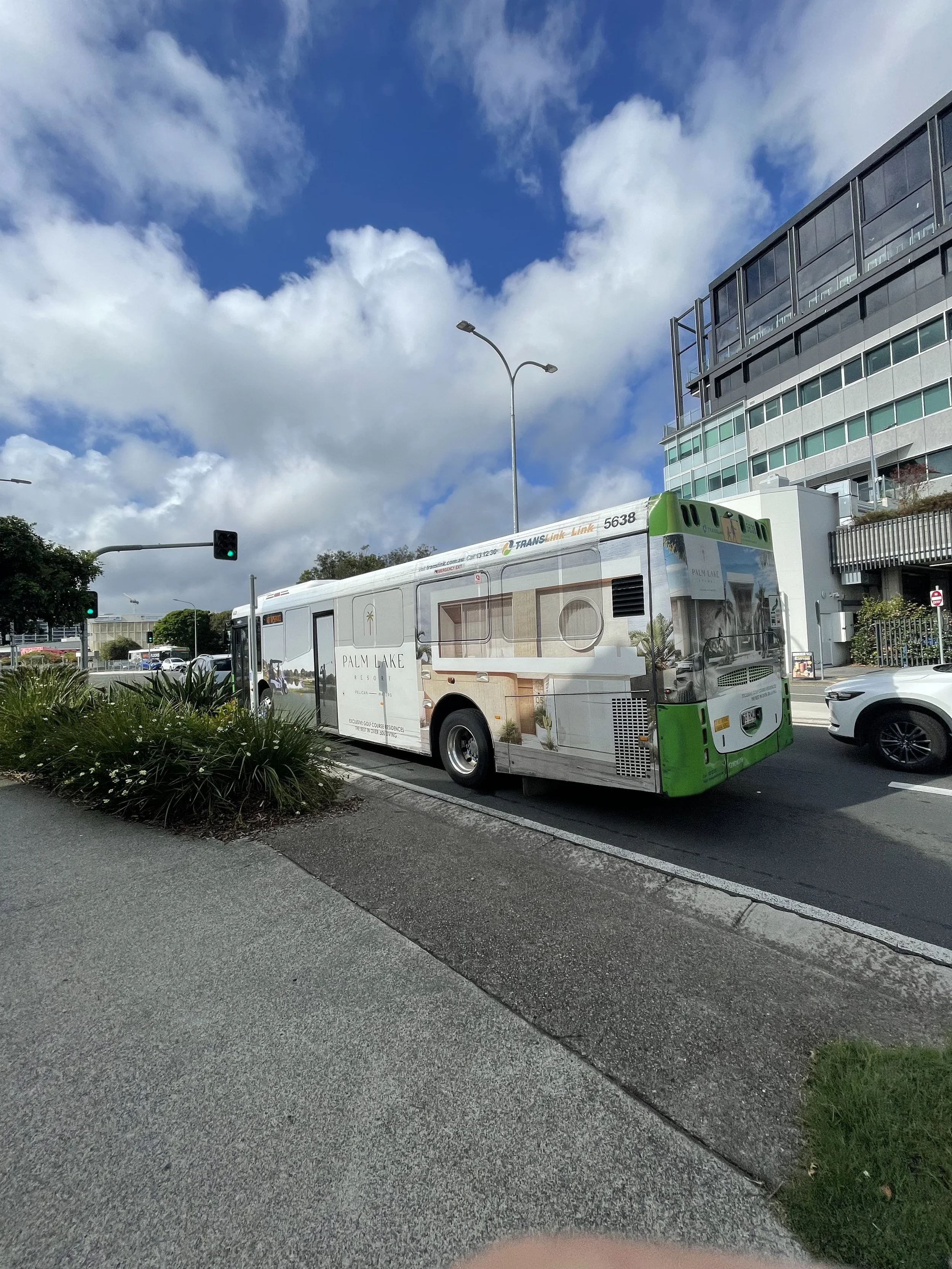 A public transit bus with a Property Developers advertisement parked at a city bus stop with green traffic lights and surrounding modern buildings and cars under a partly cloudy sky.