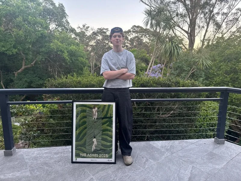 A young man with crossed arms stands on a balcony, next to a framed artwork leaning against the railing. The artwork depicts two people lying on a green, leafy garden pathway, with the text "THE ASHES 2021" at the bottom.