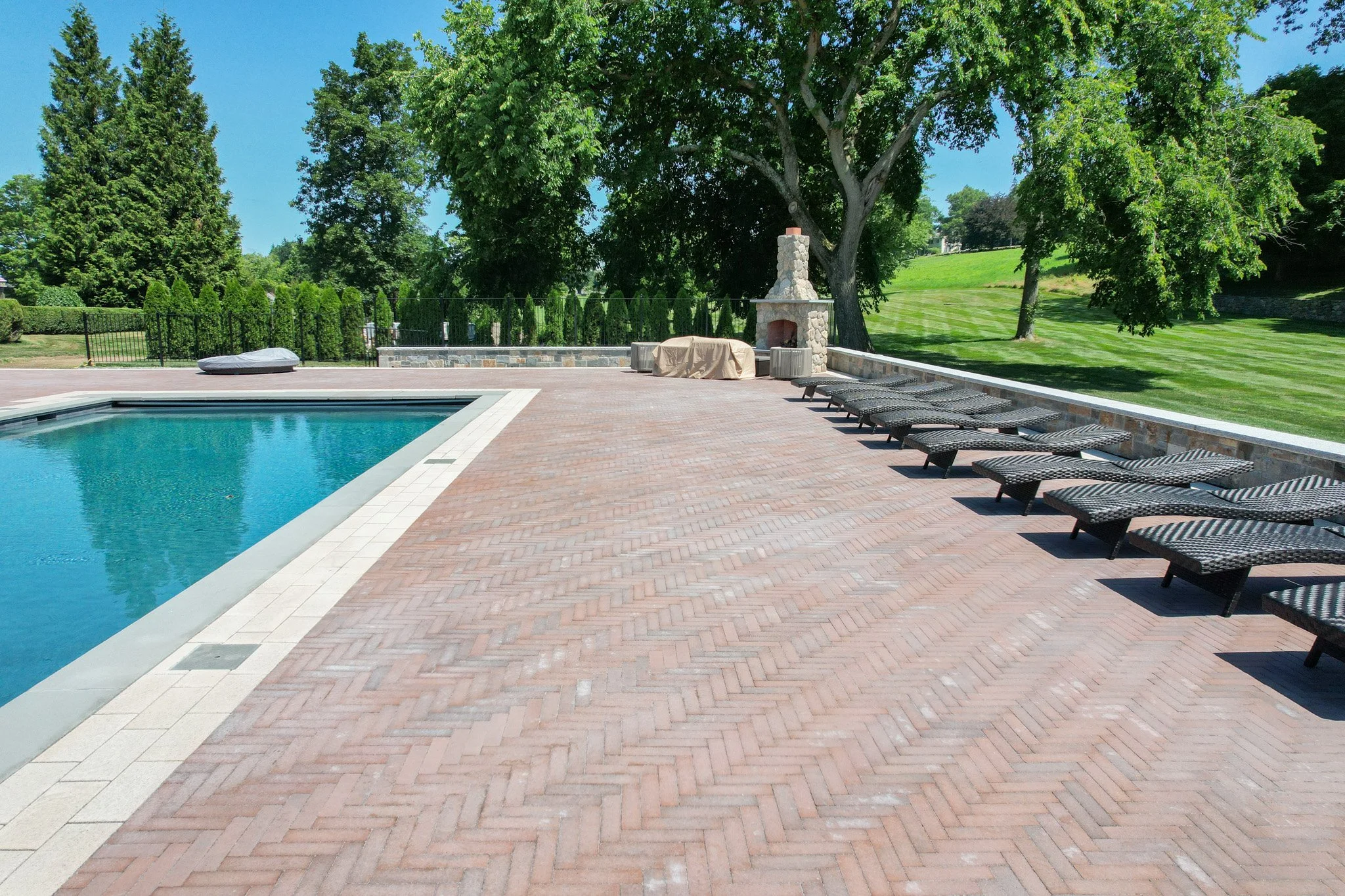 Empty swimming pool with lounge chairs, brick patio, stone fireplace, large trees, and landscaped lawn in a backyard.
