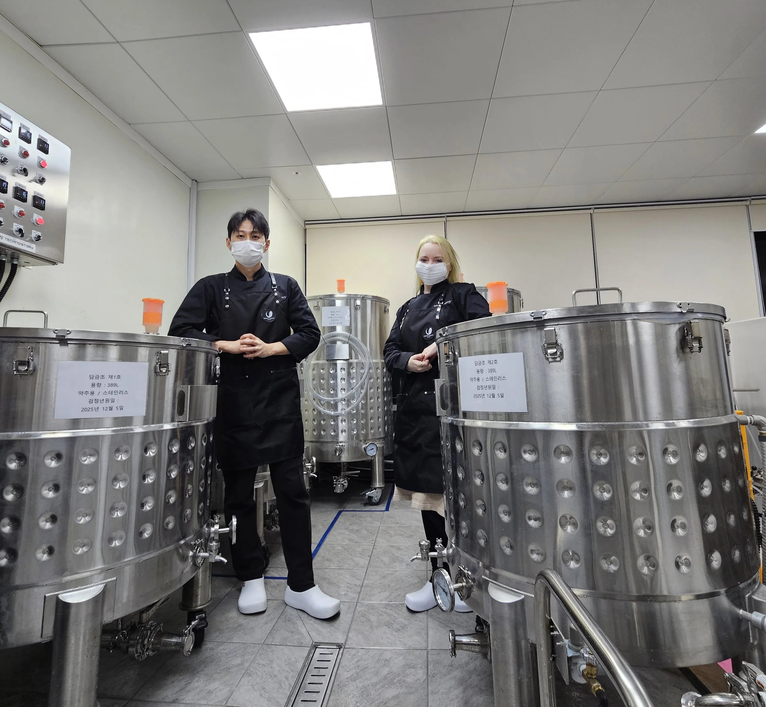 Two people in black chef uniforms and face masks stand between large stainless steel brewing tanks in a commercial kitchen or brewery. The tanks have labels, and the room is brightly lit with a tiled floor and white ceiling.
