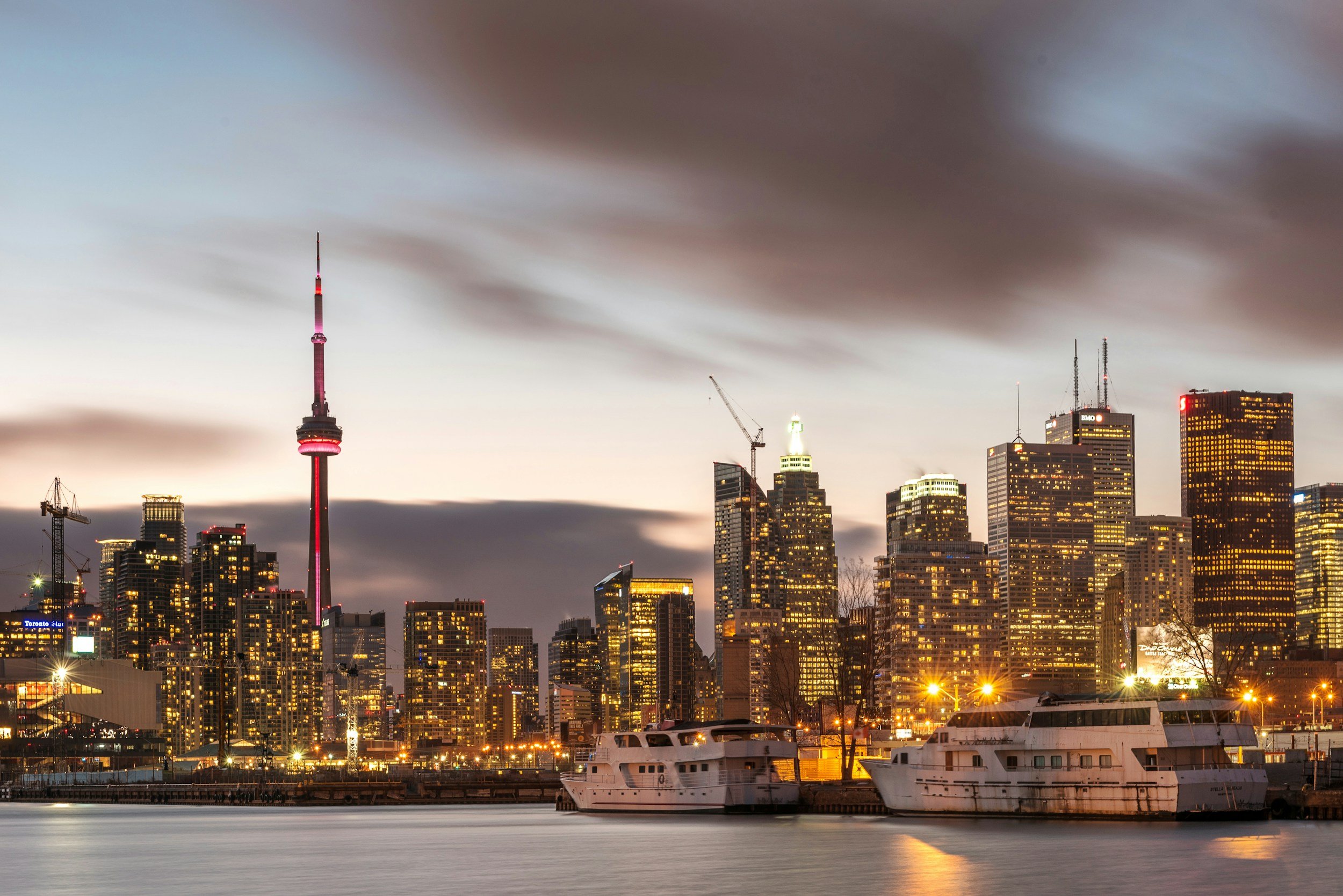 Nighttime view of Toronto city skyline with illuminated buildings, CN Tower, and boats docked on the water in the foreground.
