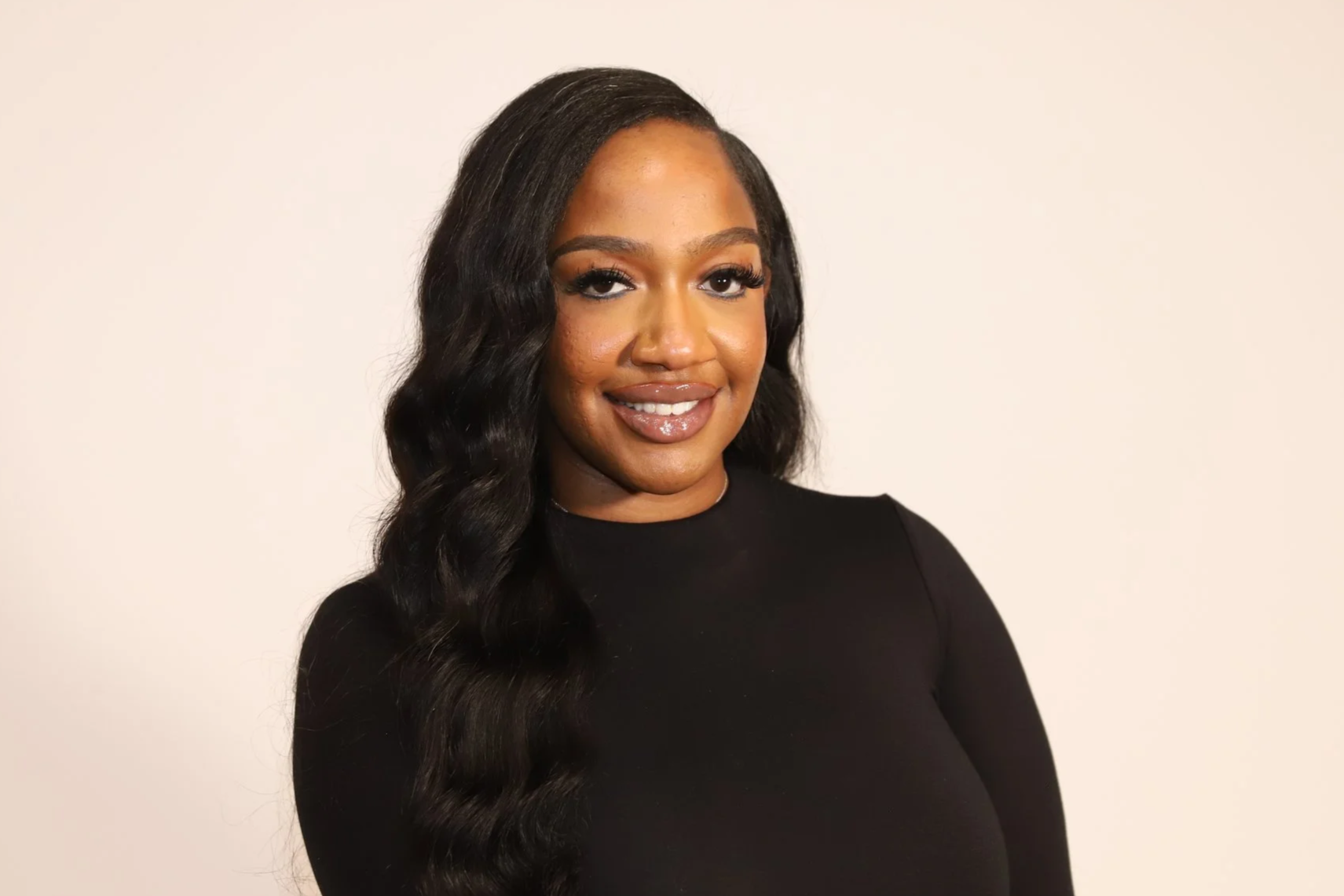 A woman with long wavy black hair wearing a black top, smiling, standing against a plain light background.