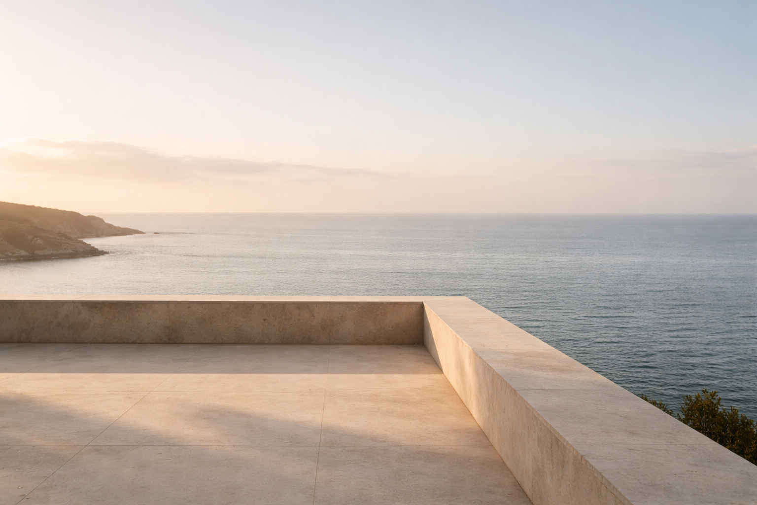 A terrace with beige stone flooring and a matching stone railing overlooking the calm ocean with a distant shoreline and a clear sky.
